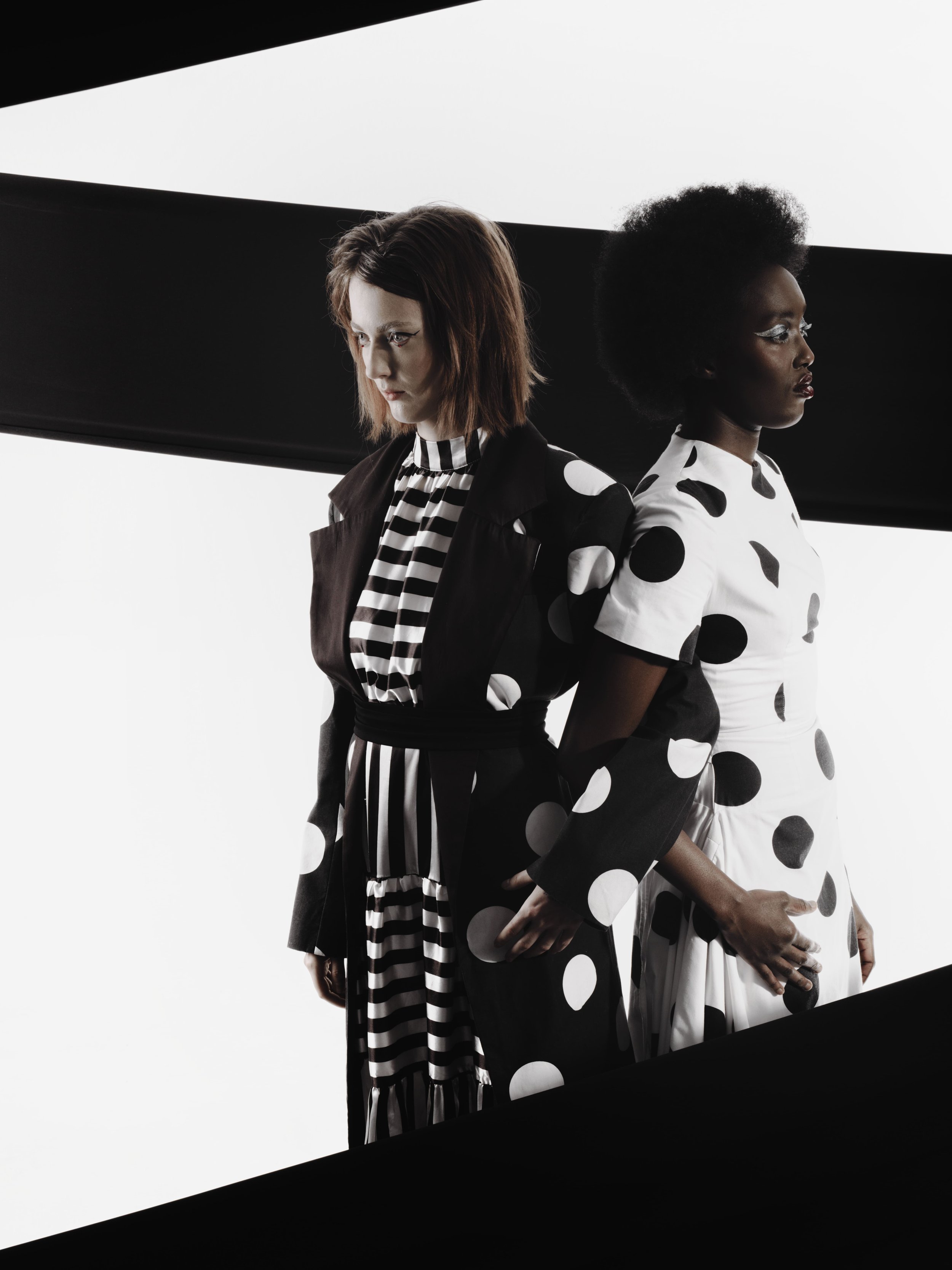 CAYLEIGH Bancroft and KIM Chakanyuka from AURA Models wearing black and white polka dot and striped clothing standing against a stark white background with black diagonal framing.