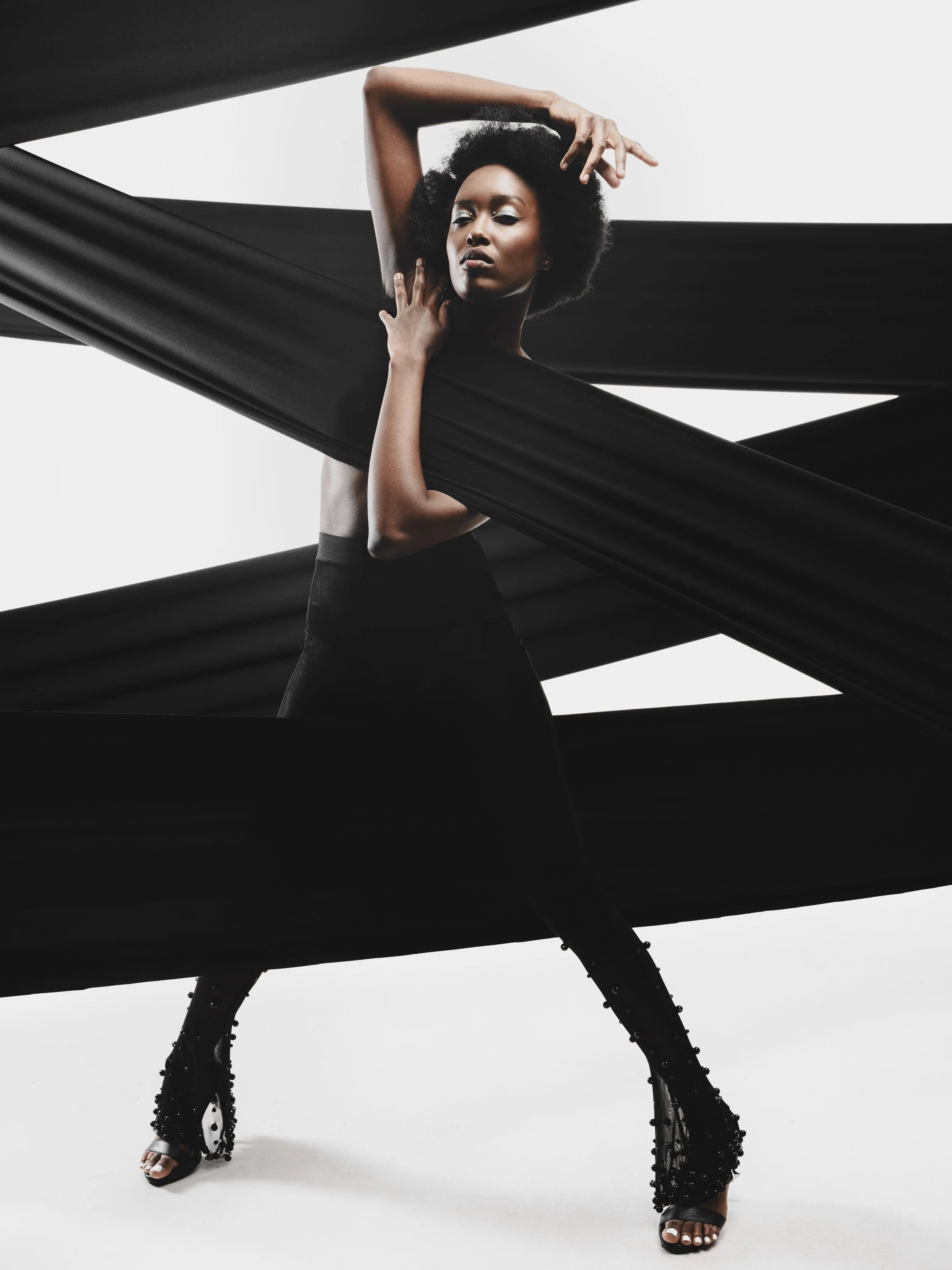 KIM Chakanyuka from AURA Models with an afro hairstyle poses with one hand on her head and the other on her neck, standing inside or near a structure shaped like an helicopter with black fabric or material draped over it.