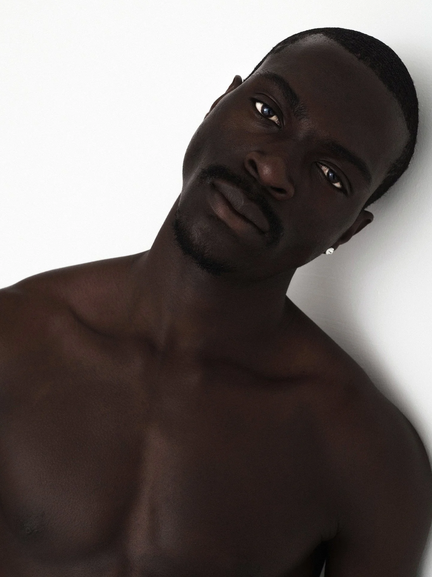 A shirtless Black man with a short haircut and a trimmed beard, leaning against a white wall, looking into the camera with a neutral expression.