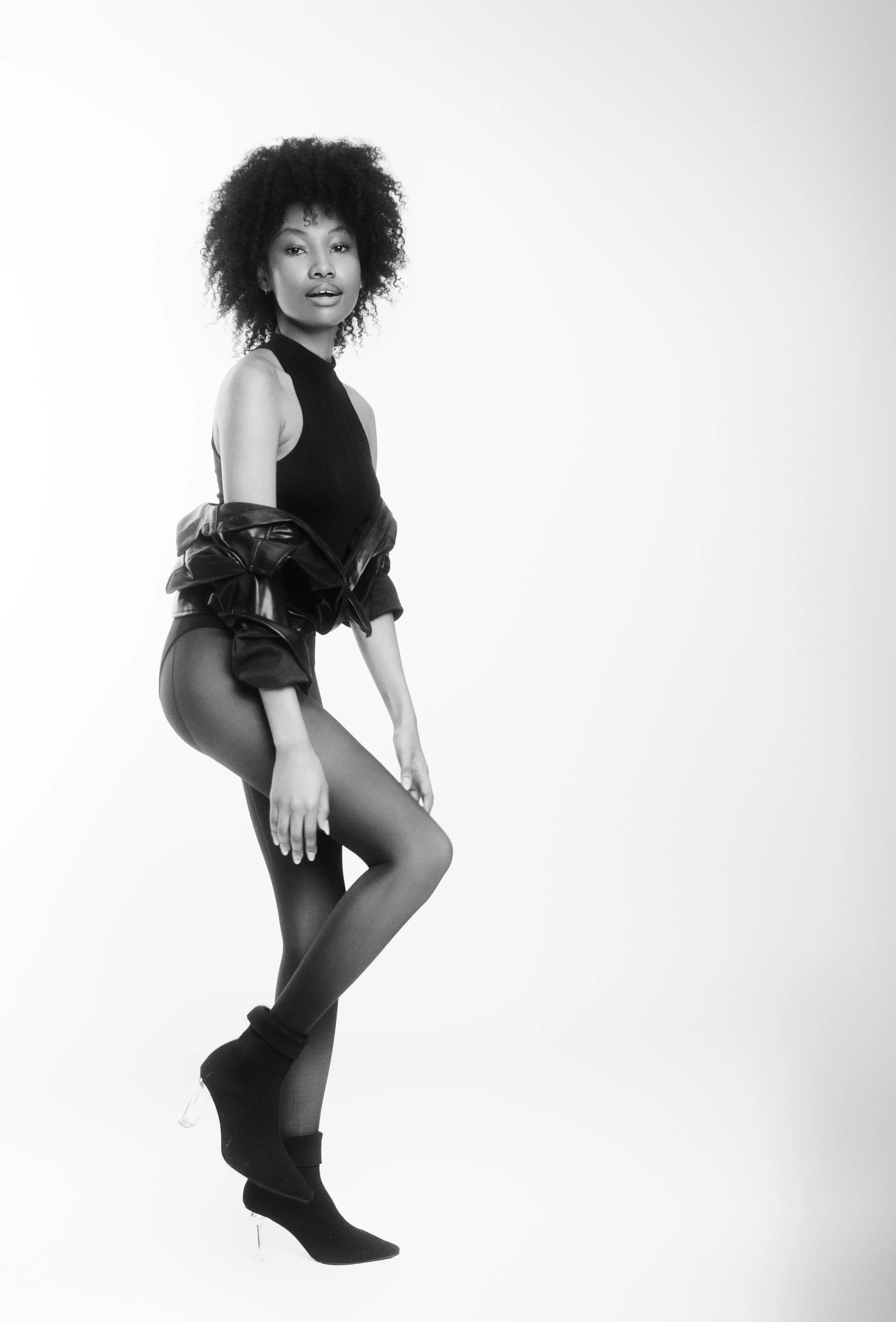 Black and white photo of QUISHA Rapoo from AURA Models with natural curly hair wearing a sleeveless top, sheer tights, and ankle boots, striking a pose against a plain white background.