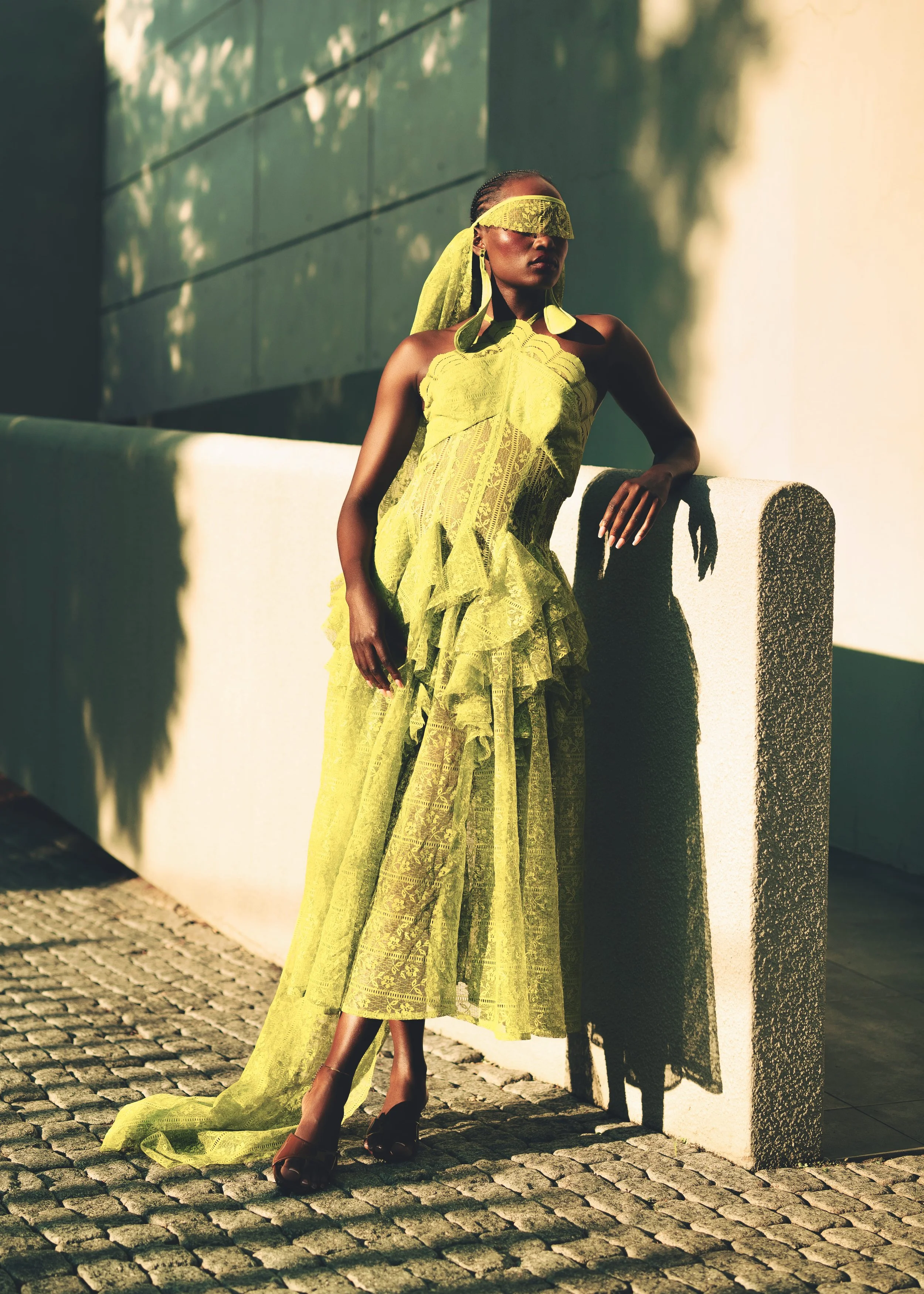 SHALOM Musvosvi from AURA Models wearing a bright yellow lace dress with a matching veil over her eyes, standing outside against a light gray wall with shadows from trees, on a cobblestone street, with one hand resting on the wall. 