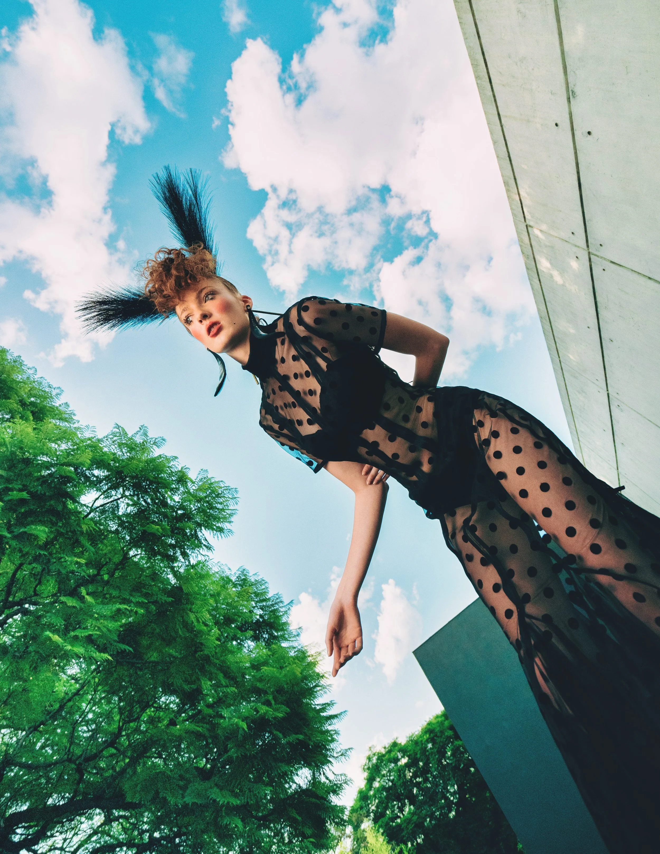 CAYLEIGH Bancroft from AURA Models with short curly hair and wearing a black polka dot sheer dress is looking over her shoulder outdoors on a bright day with blue sky and white clouds, surrounded by green trees, with a gray building in the background
