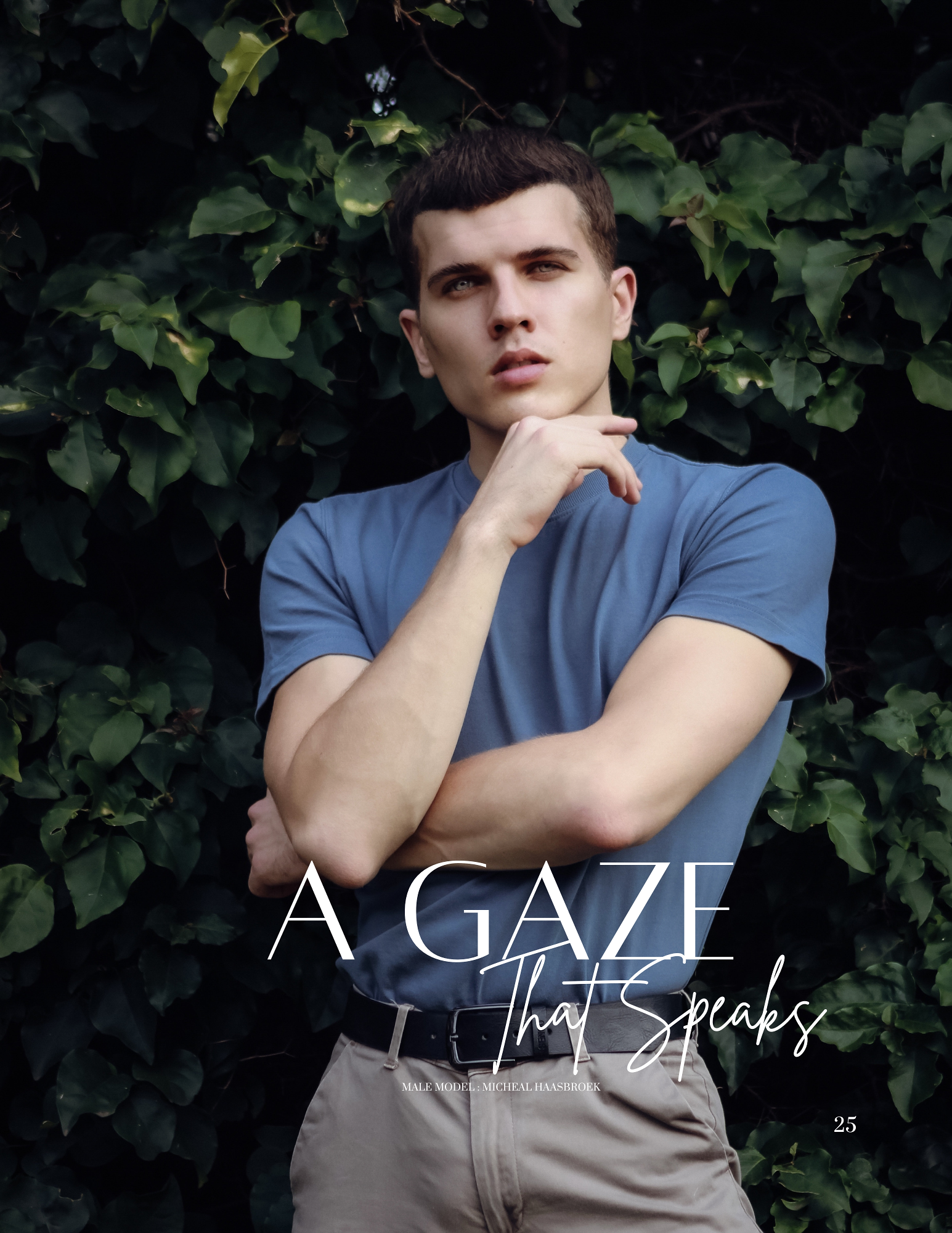 A young man with short dark hair, wearing a blue t-shirt, standing in front of green leafy bushes, looking thoughtfully at the camera with his right hand resting on his chin.