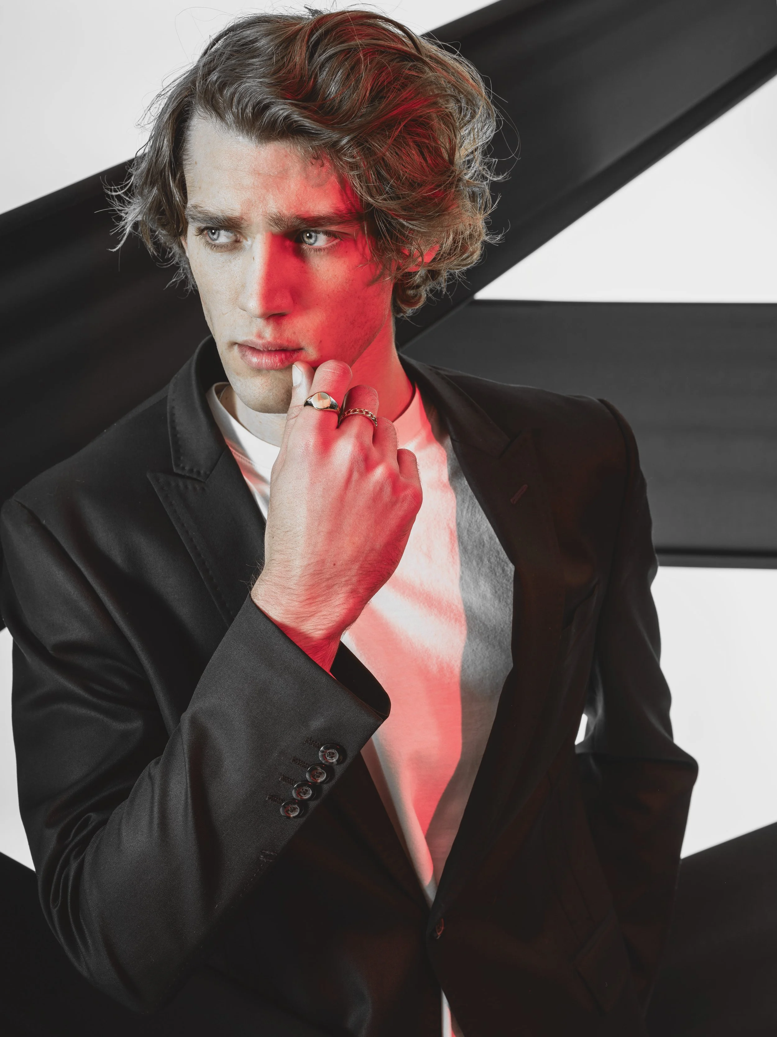 JOSH Gradwell in a black blazer over a white shirt, standing against a black and white background. He has a contemplative expression, resting his chin on his hand with rings on his fingers, and the image has a red tint on the left side of his face.