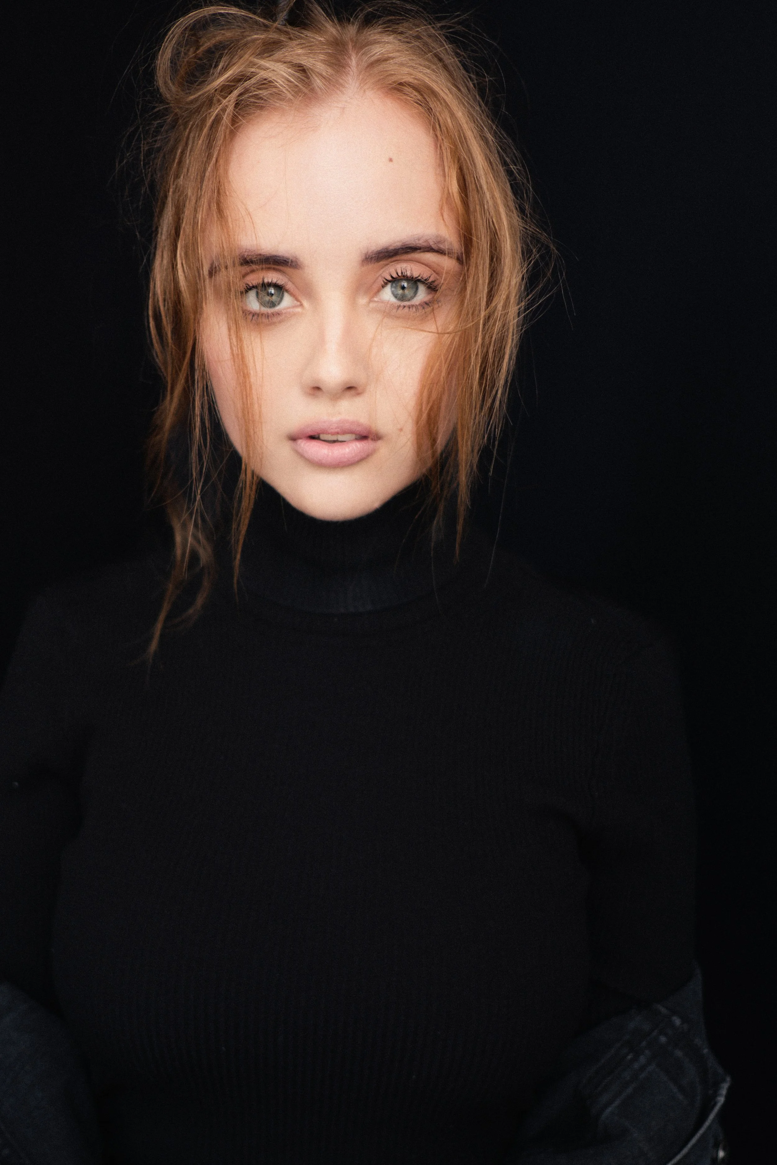 Portrait of DANIELLE Laubscher from AURA Models with red hair, blue eyes, wearing a black turtleneck, against a dark background.