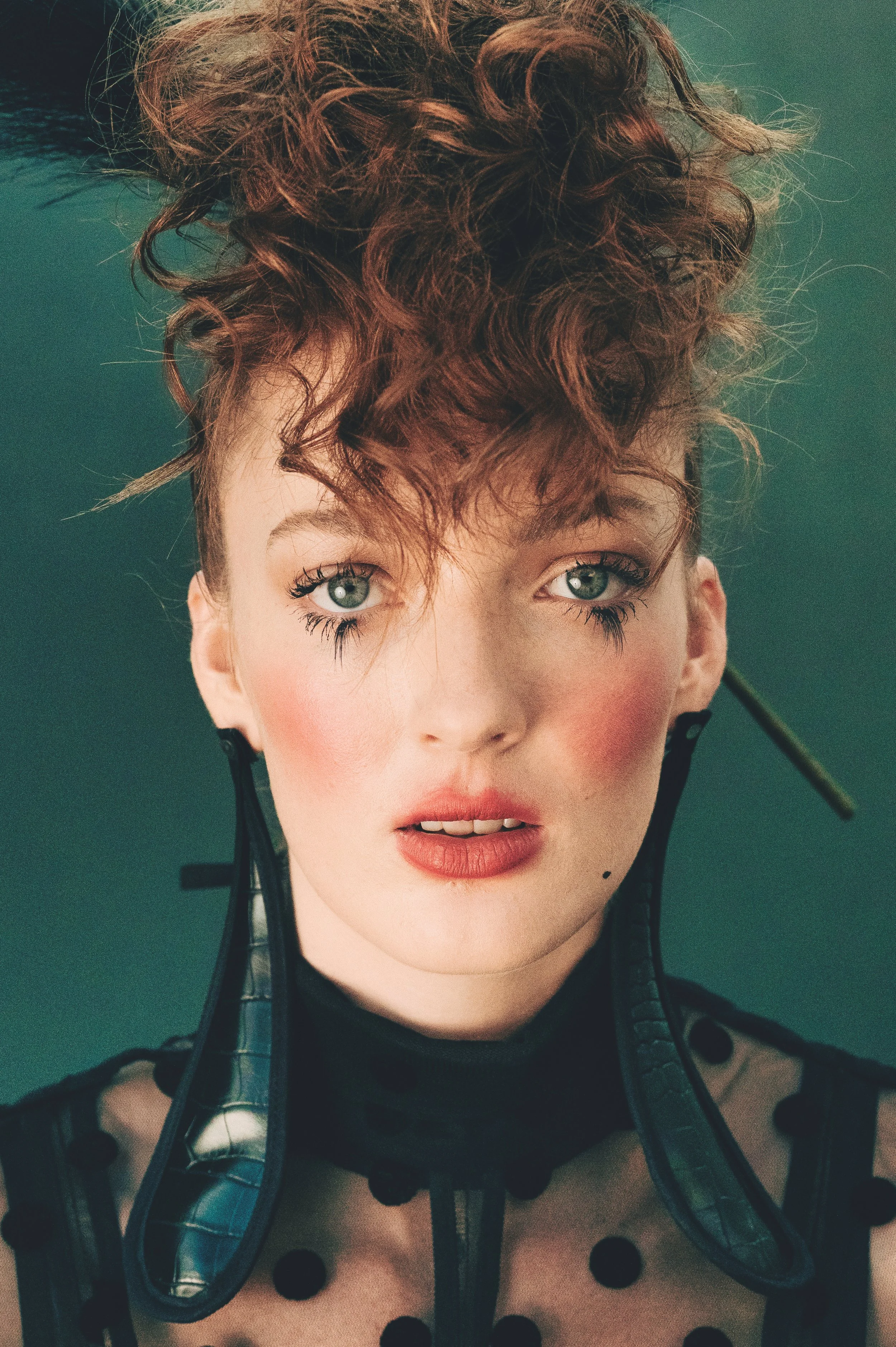 CAYLEIGH Bancroft from AURA Models with curly red hair and blue eyes wearing a sheer polka dot top and a black leather harness with straps.