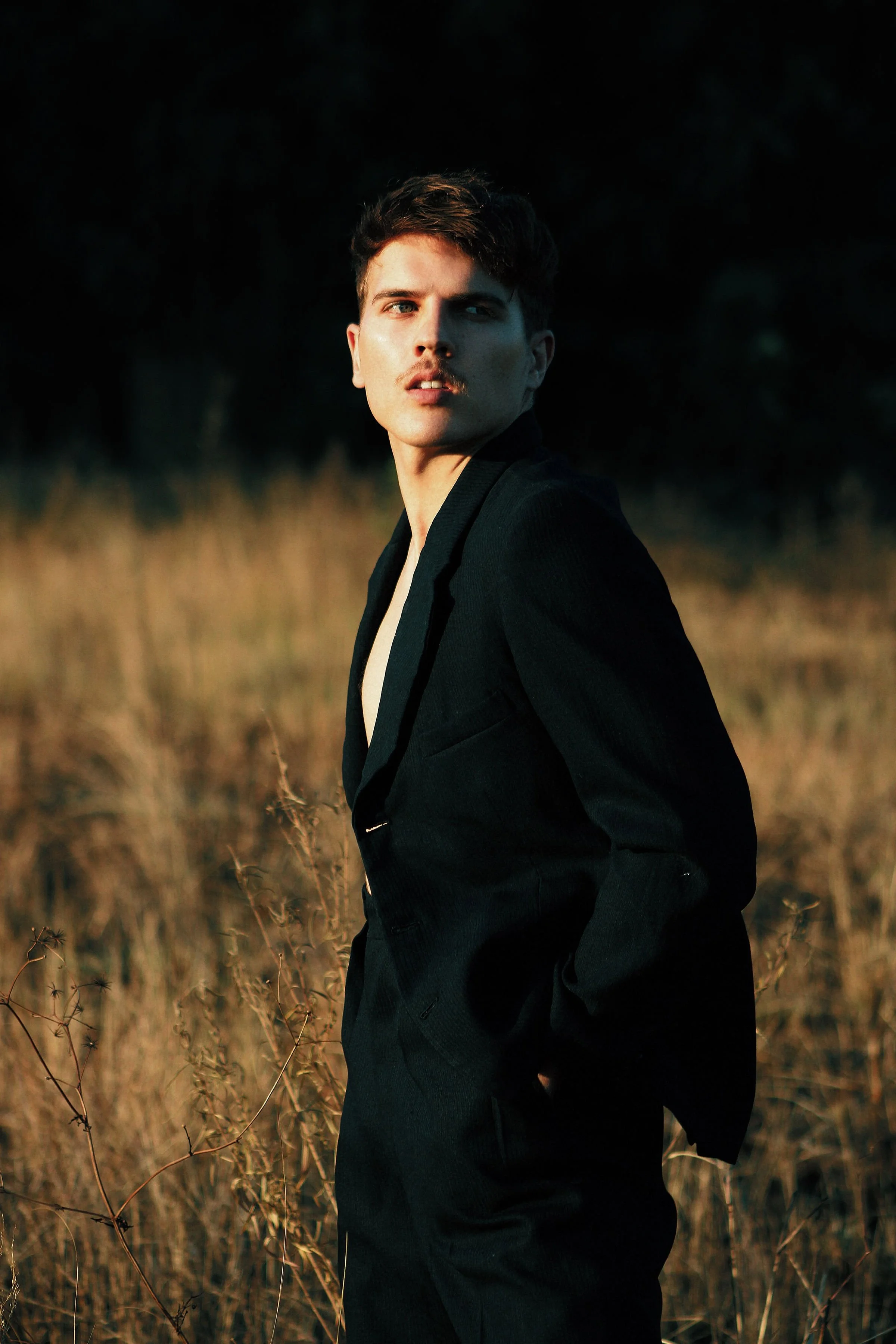 A young man standing outdoors in a field of tall, dry grass at sunset, wearing a black blazer with hands in pockets, looking to the side.