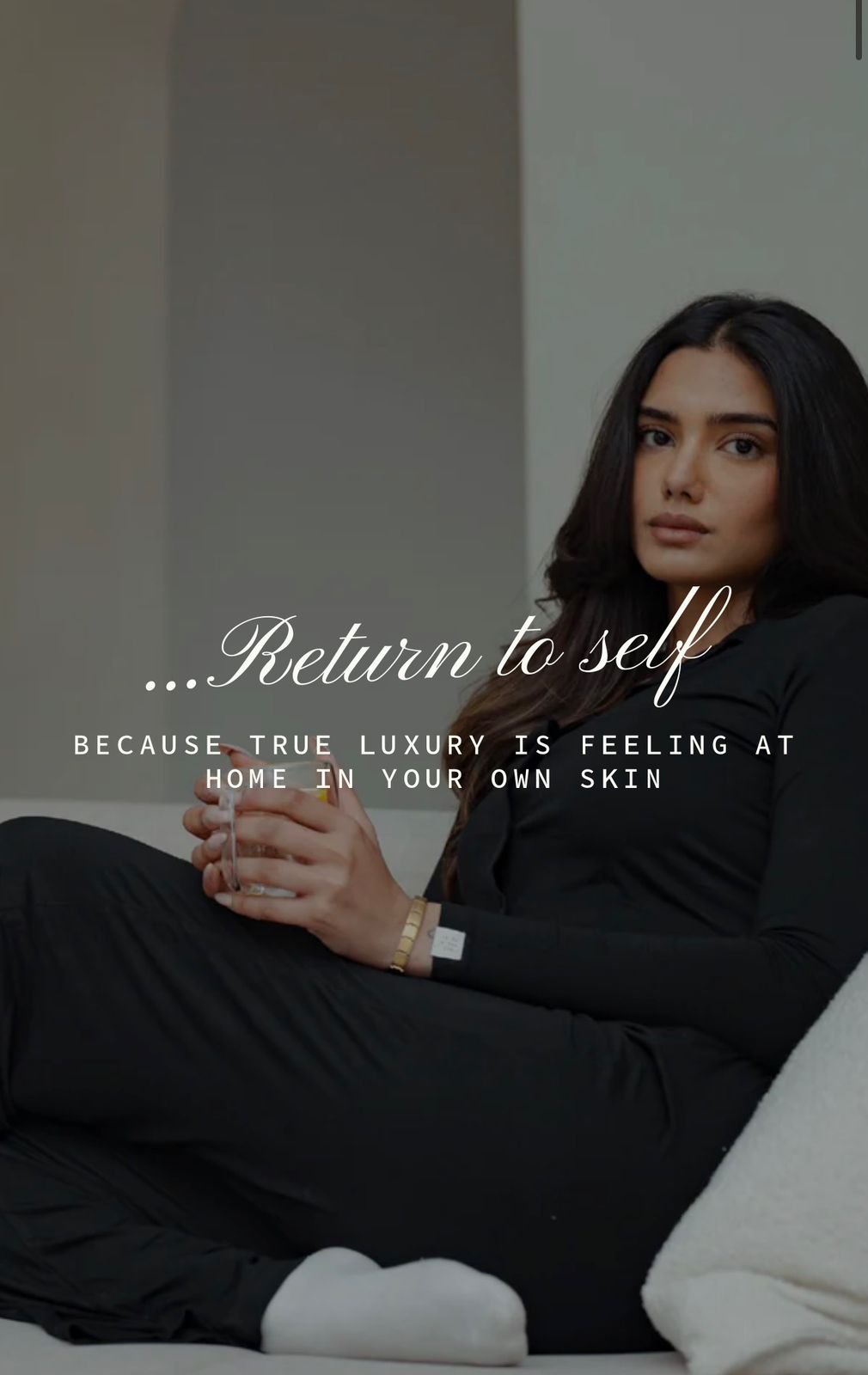 EMILY Francis from AURA Models with long dark hair sitting on a sofa holding a mug, wearing a black outfit, with a text overlay that reads 'Return to self because true luxury is feeling at home in your own skin'.