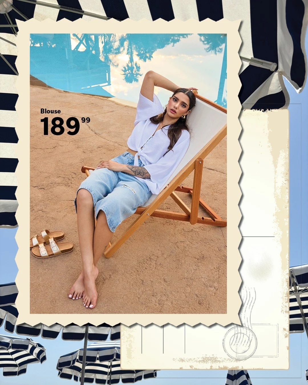 A woman with long dark hair, wearing a white tied blouse and distressed denim shorts, is sitting on a wooden lounge chair at the beach. She has one arm behind her head and the other resting on her lap, with sandals on the sand nearby. The background 