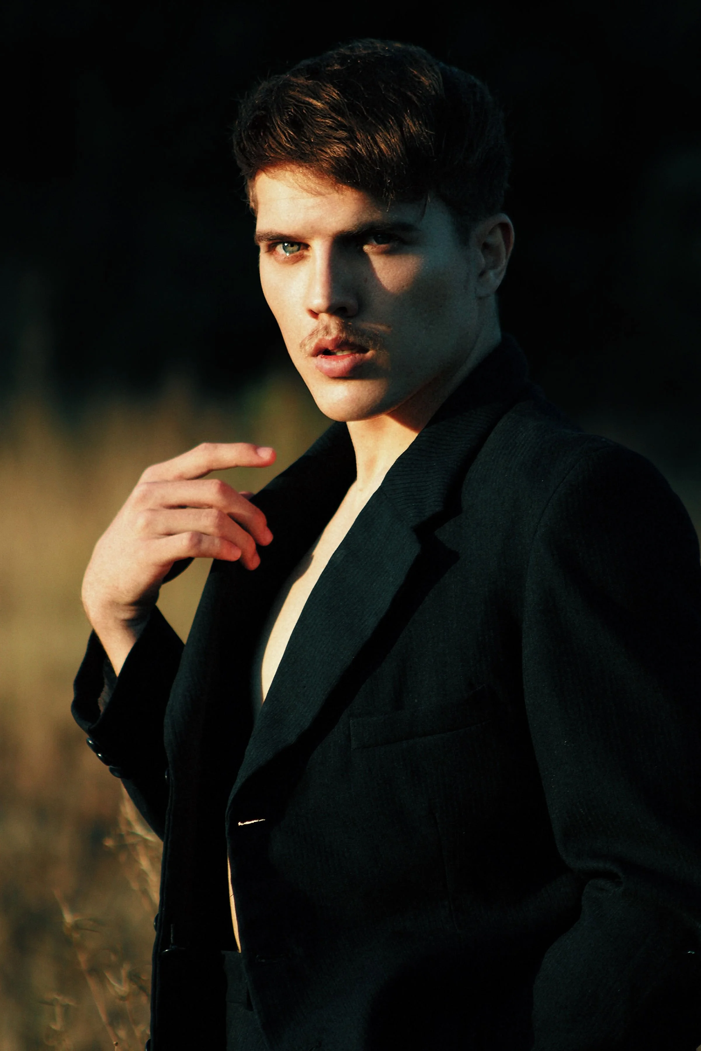 Young man with light skin and short, styled dark hair, wearing a black blazer, posing outdoors at sunset or dusk with a serious expression.