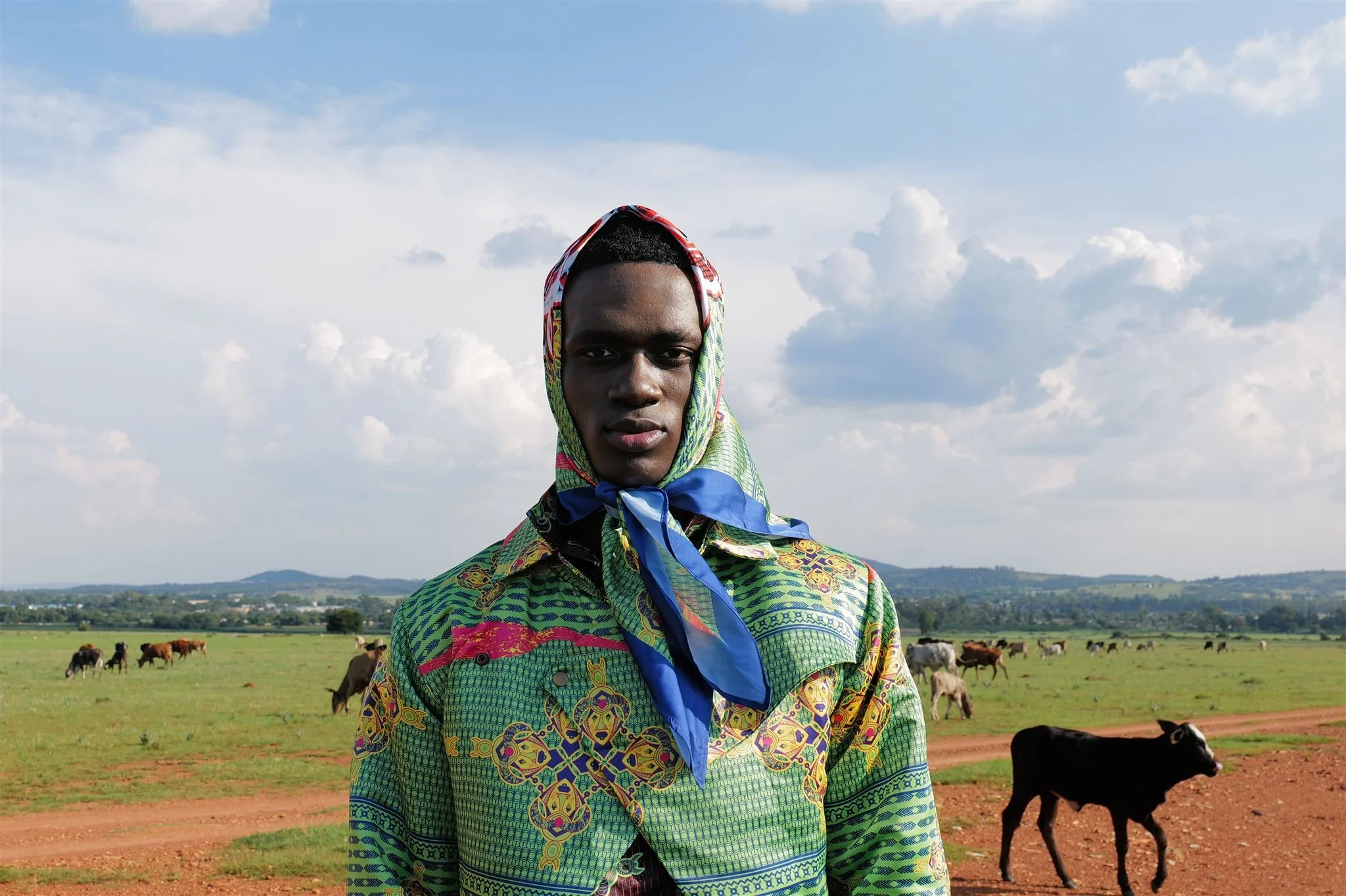 MBHURI Baloyi from  wearing a vibrant, patterned shirt and a colorful hooded scarf with a blue ribbon tied at the neck, standing outdoors in a rural landscape with cows grazing in the background and mountains on the horizon under a partly cloudy sky.