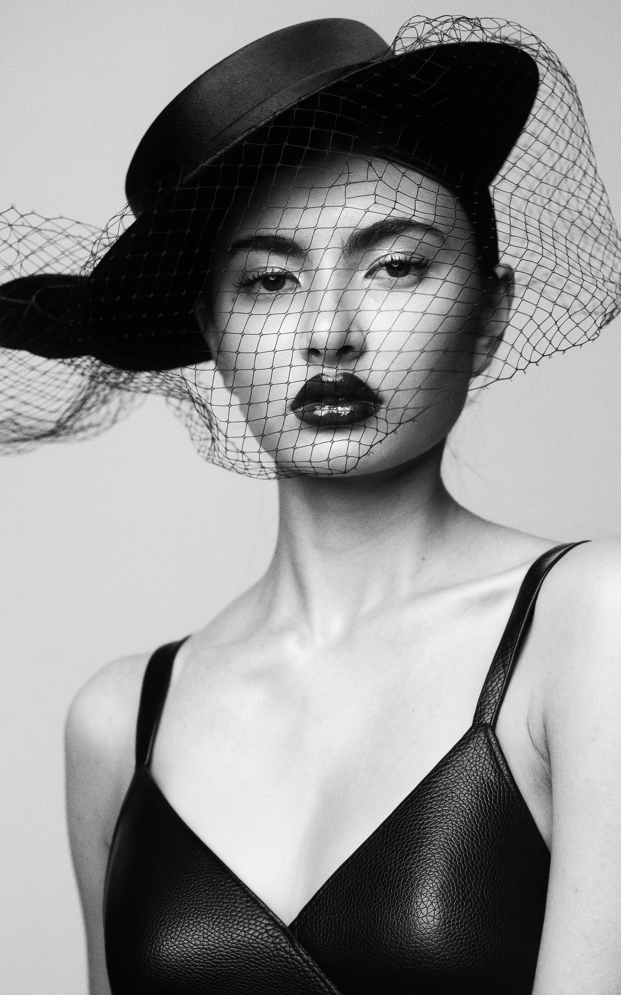 Black and white portrait of KEIRA Louw from AURA Models wearing a black leather top and a hat with a net veil covering part of her face.