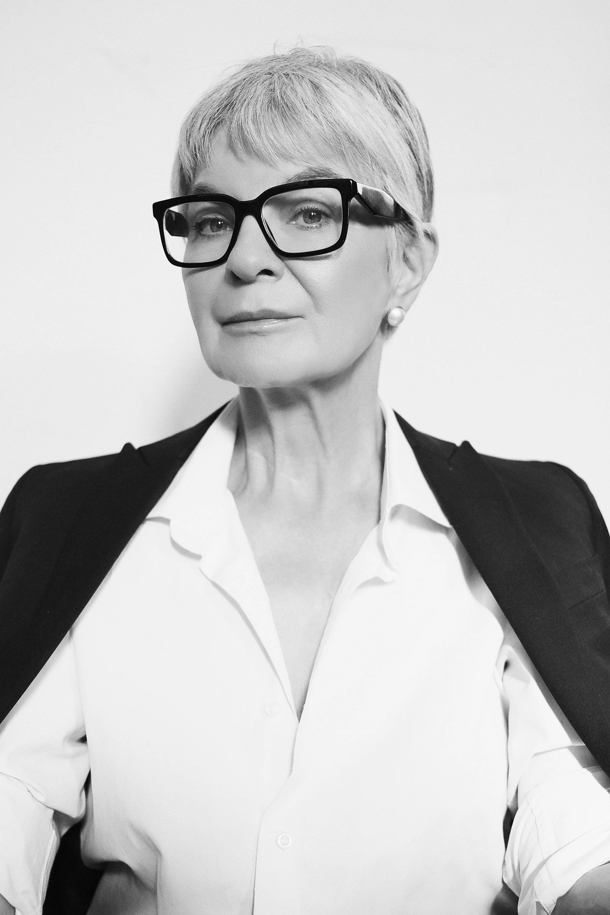 A black-and-white portrait of BRIDGET van Zyl from AURA Models with short hair, wearing large glasses, pearl earrings, a white button-up shirt, and a dark blazer draped over her shoulders.