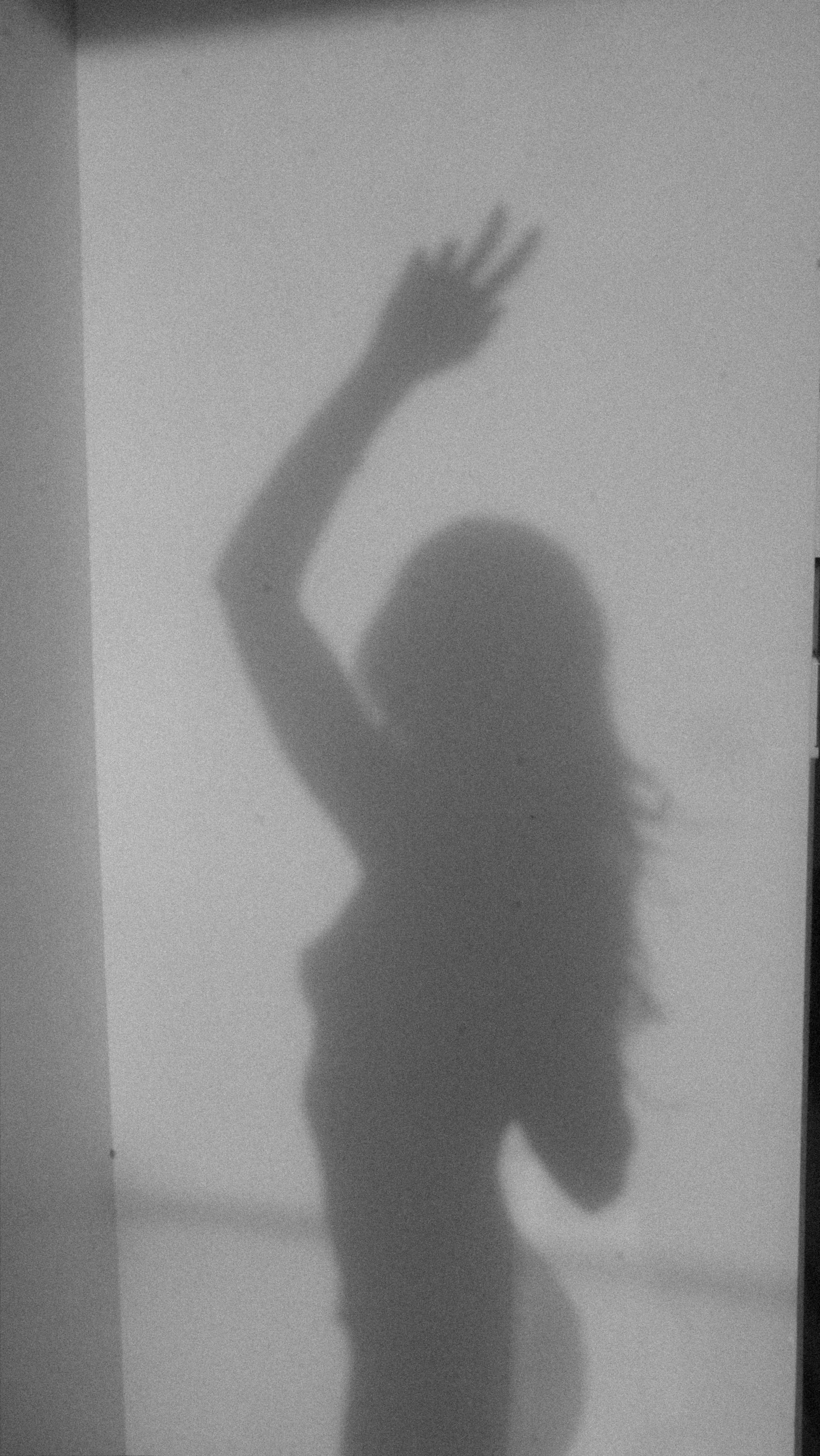 Shadow of a person, possibly a woman, with long hair, raising one arm with fingers splayed on a plain wall.