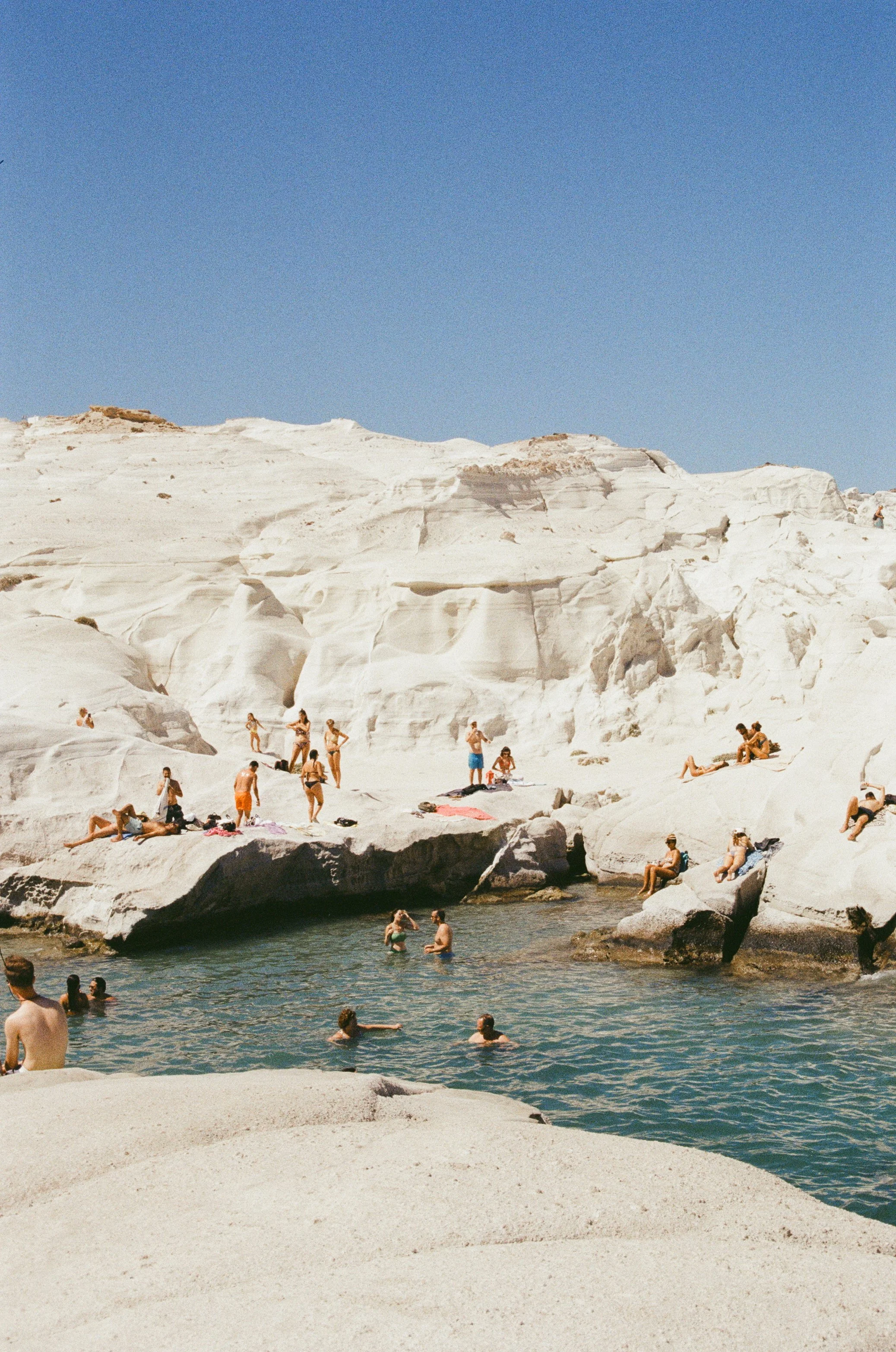 People swimming and sunbathing on white rocks by the water under a clear blue sky.