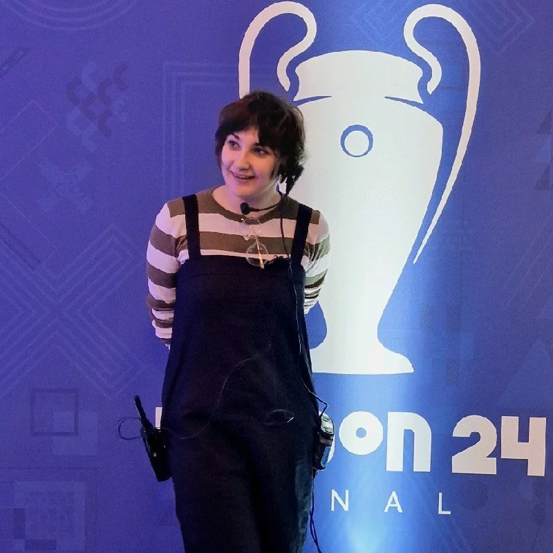 Woman with short dark hair wearing a striped long-sleeve shirt and black overalls standing in front of a blue backdrop with a stylized trophy logo and the text "on 24 NATIONAL."