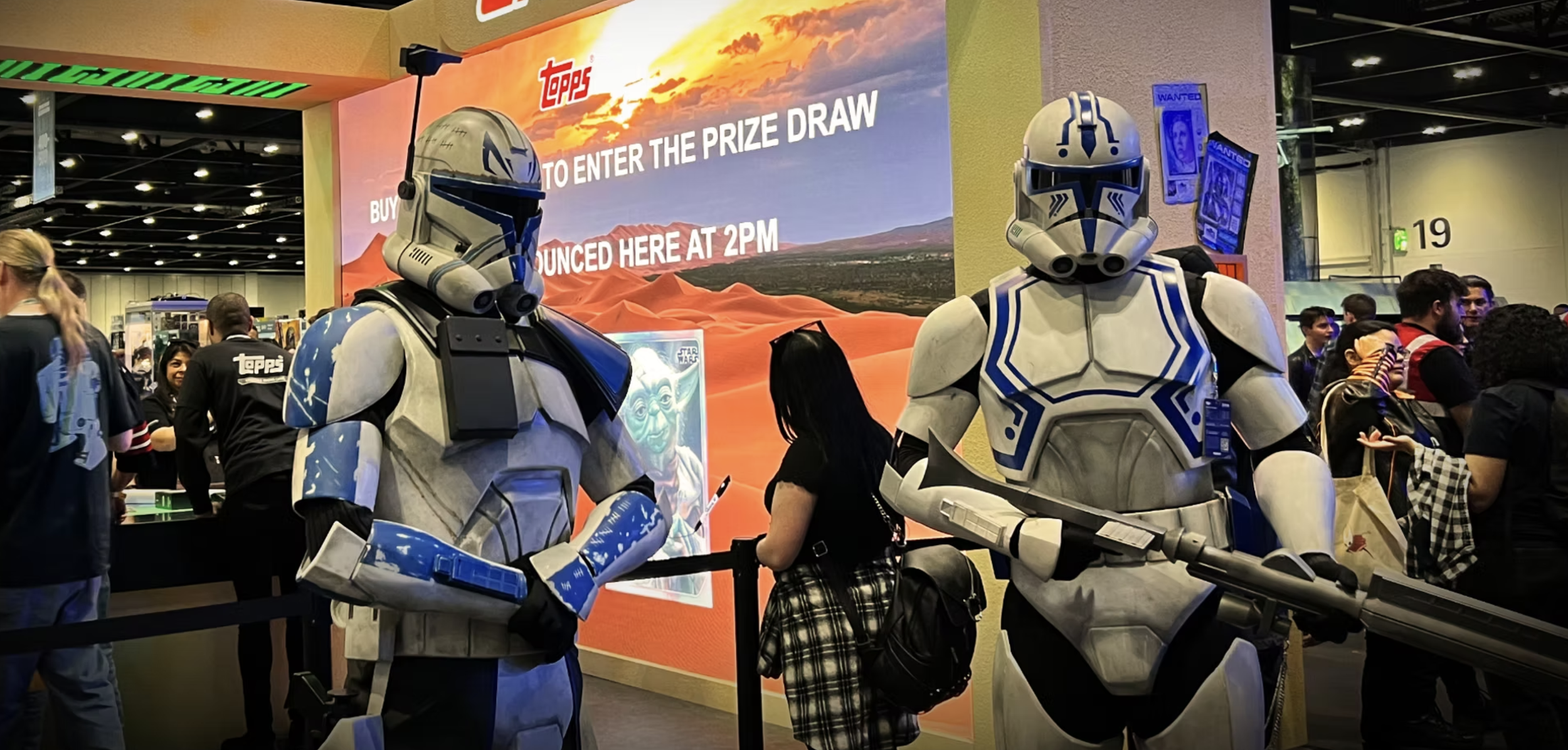 Two individuals dressed as clone troopers from Star Wars at a convention, standing in front of a promotional booth with a large screen behind them displaying the Topps logo and event details, while a crowd of people is visible in the background.
