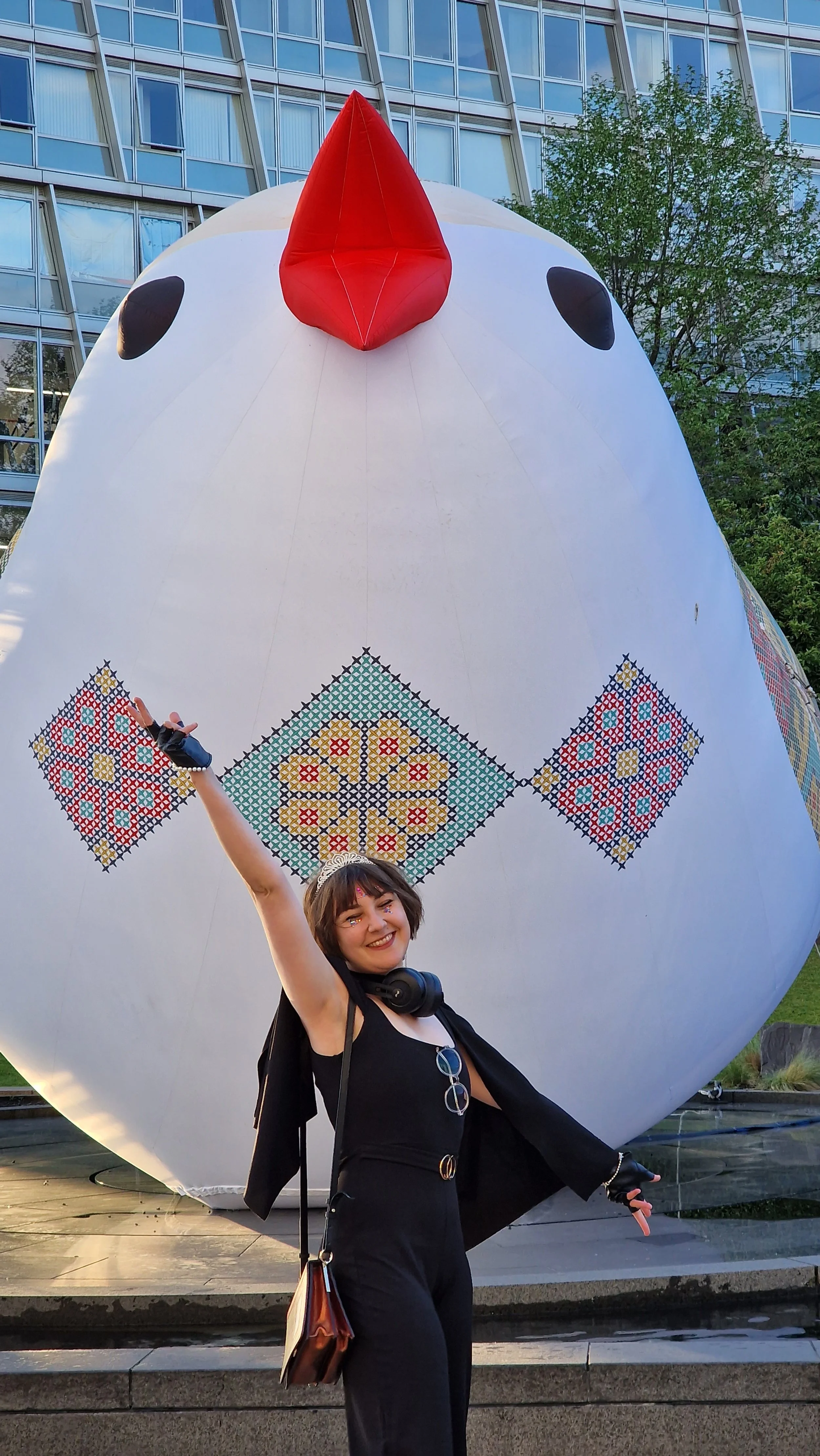 A smiling woman in black outfit with glasses hanging from her neck, standing outdoors in front of a large inflatable chick sculpture with a red beak and black eyes, decorated with colorful embroidered patterns.