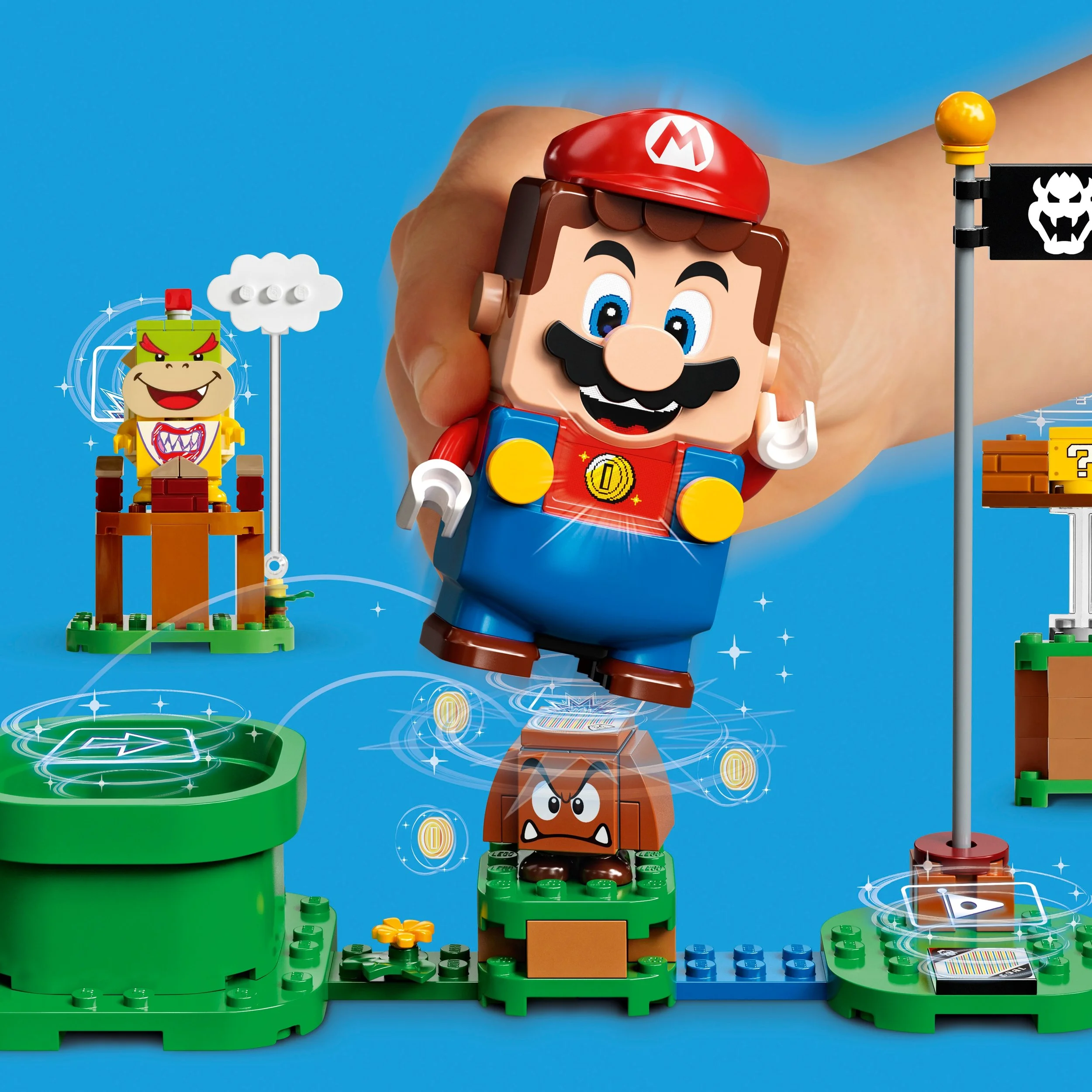 A large hand holding a Mario LEGO figure from the Super Mario series, surrounded by other LEGO Super Mario elements, including Bowser, a flag with a Koopa Troopa shell, and a green pipe, set against a bright blue background.