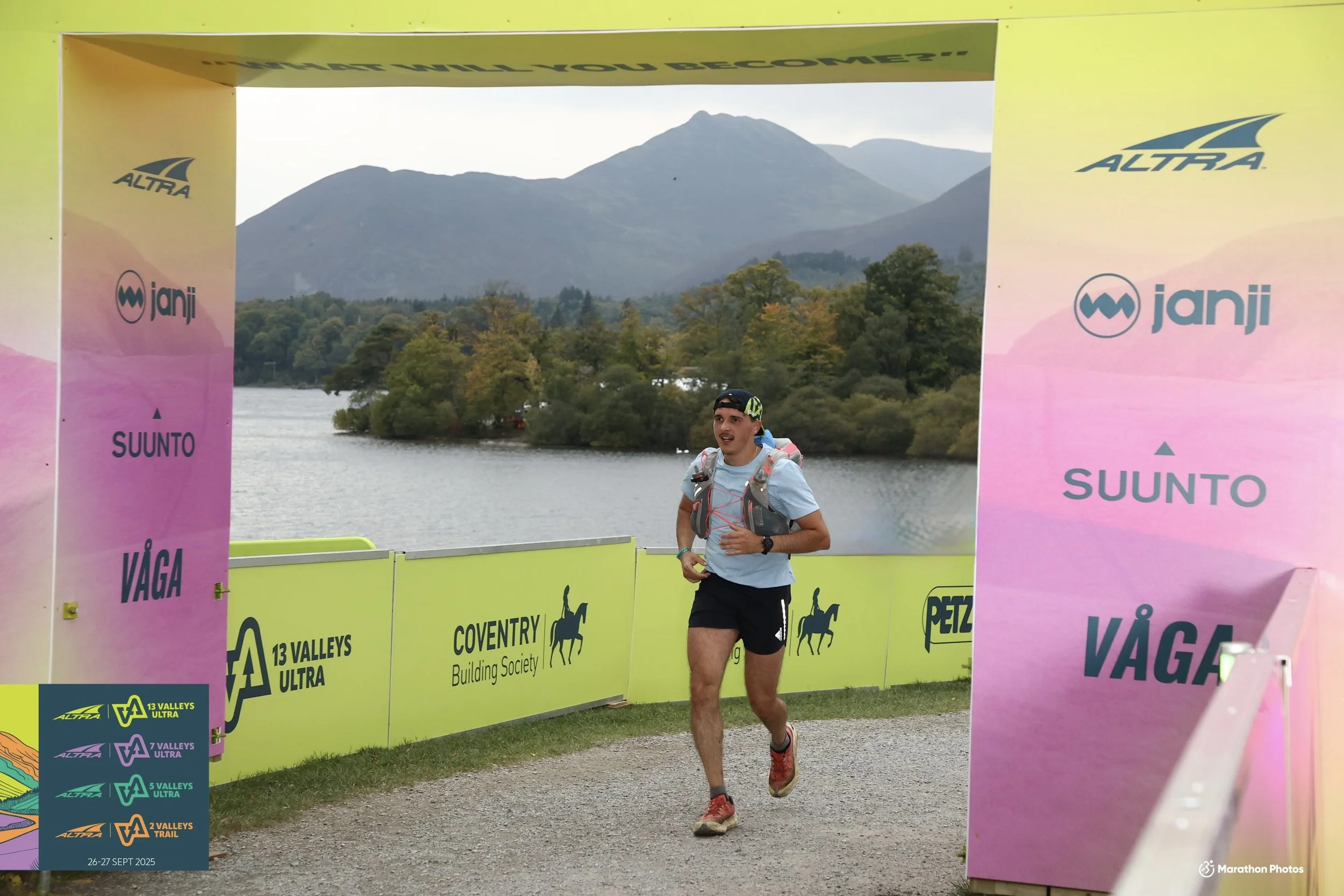 Runner crossing finish line at 13 Valleys Ultra race, with mountains, lake, and trees in background, during daytime.