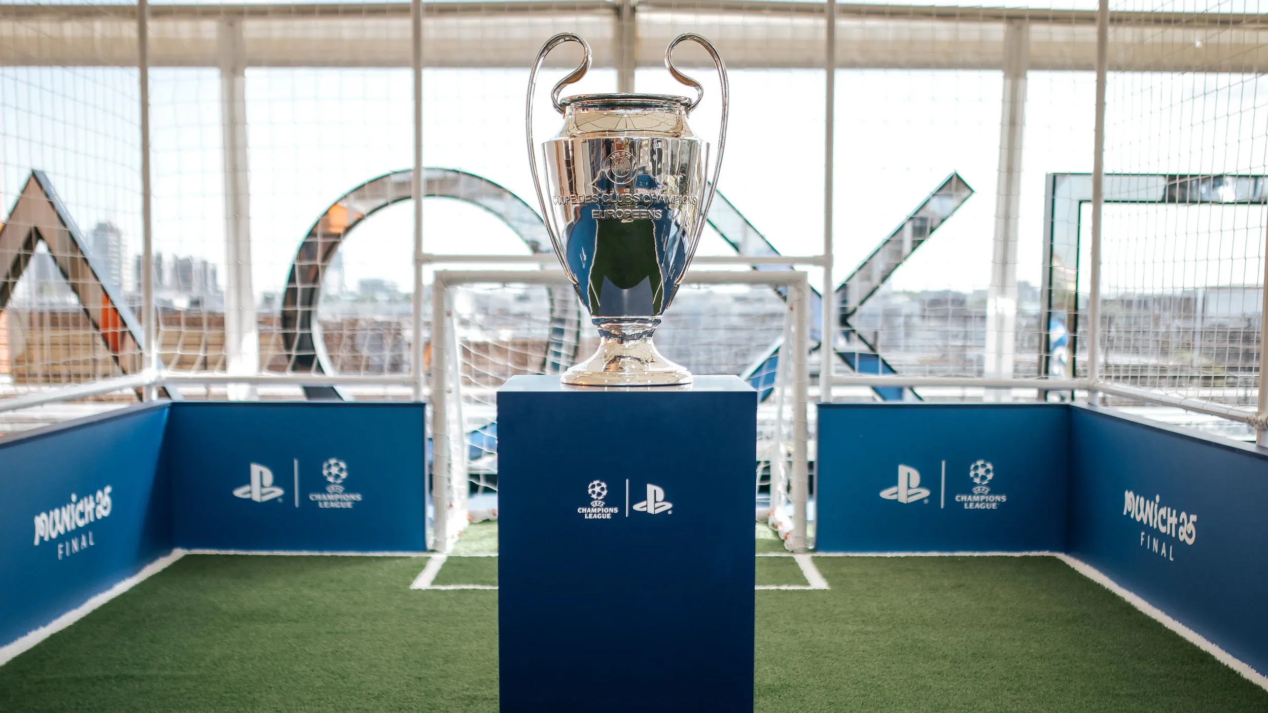 The UEFA Champions League trophy displayed on a blue pedestal inside a small football field with a goal and blue banners, celebrating the final match with the Champions League logo.