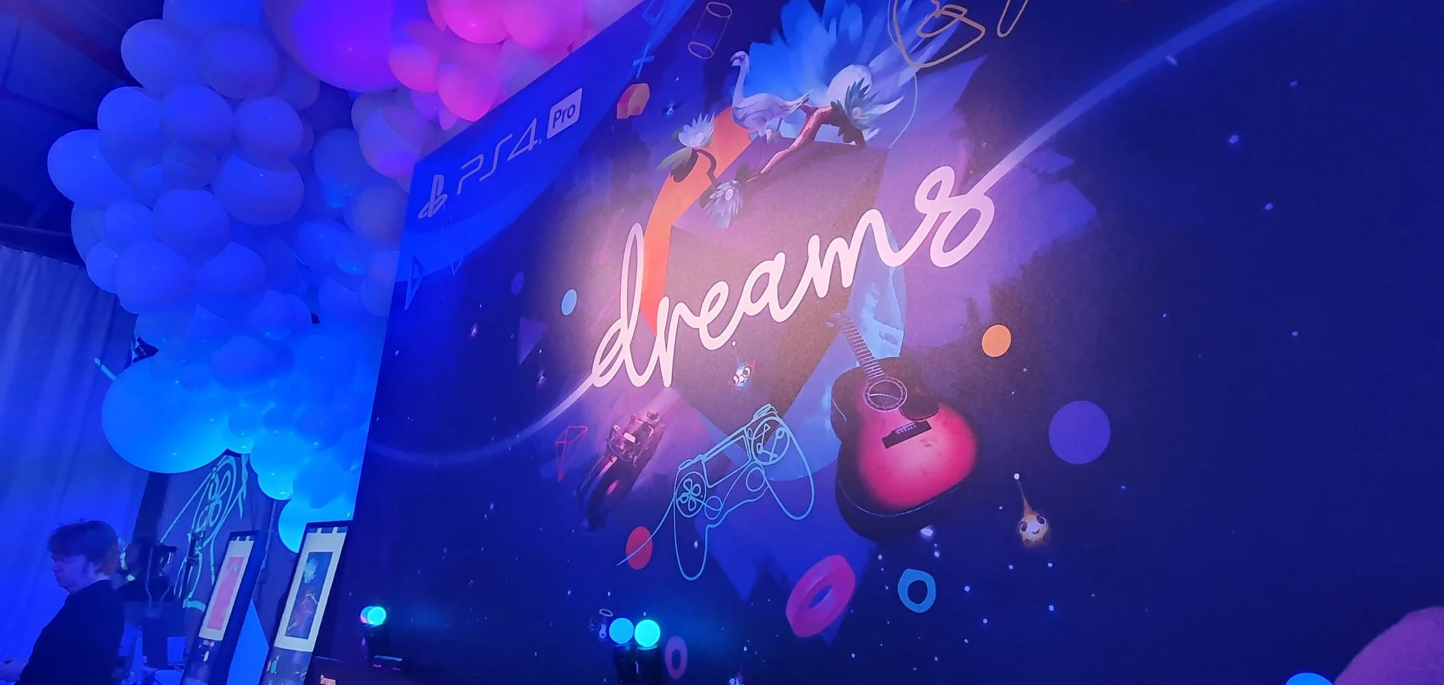 A large banner displays the word 'dream' with colorful gaming and musical icons, such as a guitar, game controller, and balloons, in a vibrant, space-themed setting at a gaming event.