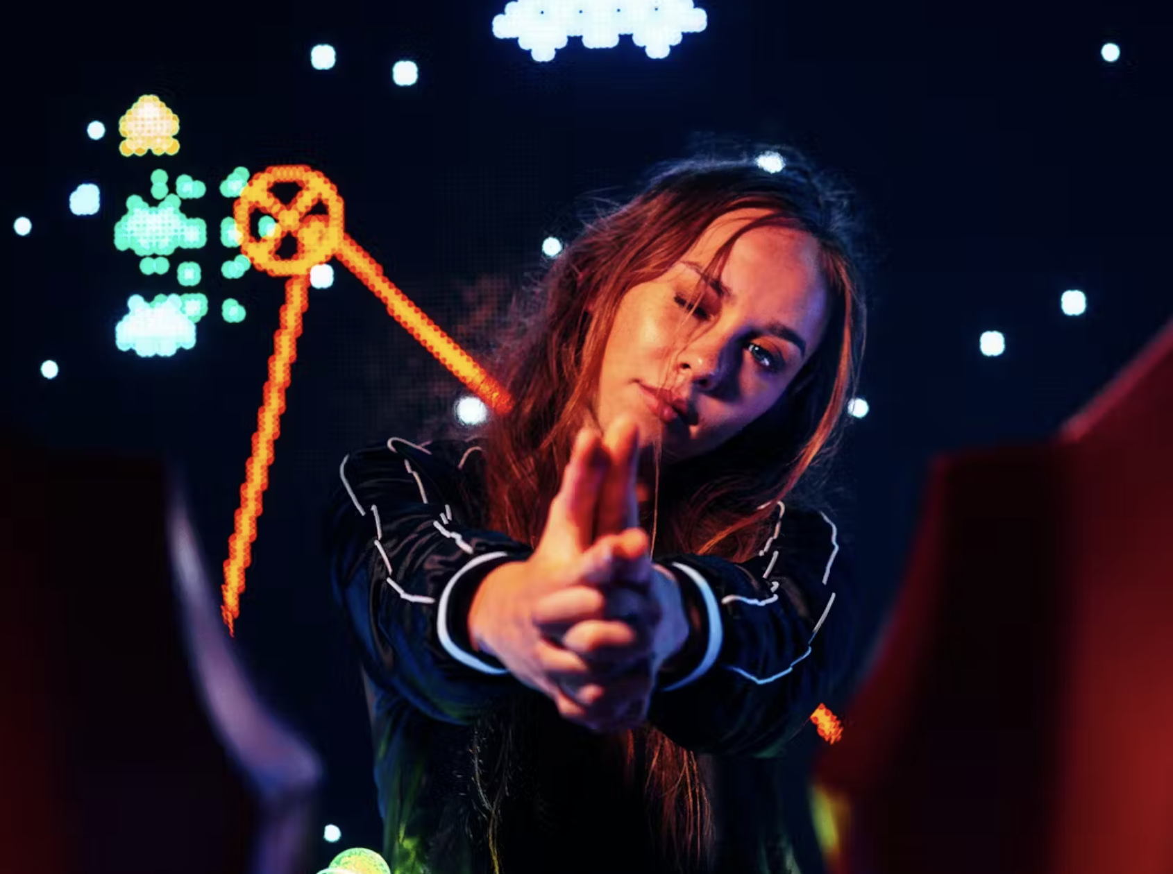 A young woman with long hair, wearing a black jacket with white piping, points towards the camera with her right hand in front of a background of neon lights and pixel art of clouds, stars, and a red spaceship in a dark setting.