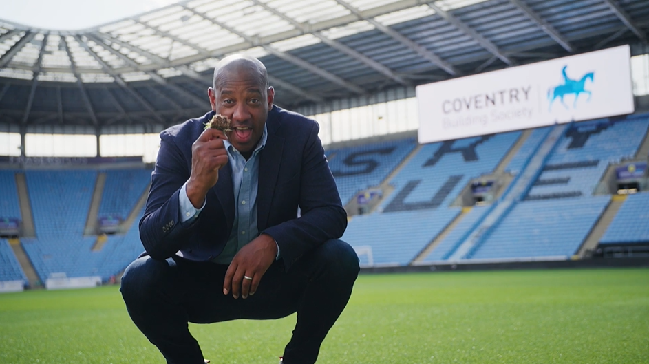 Man in business suit squatting on a football field, smiling and holding a snack, stadium seats and a Coventry Building Society billboard in the background.