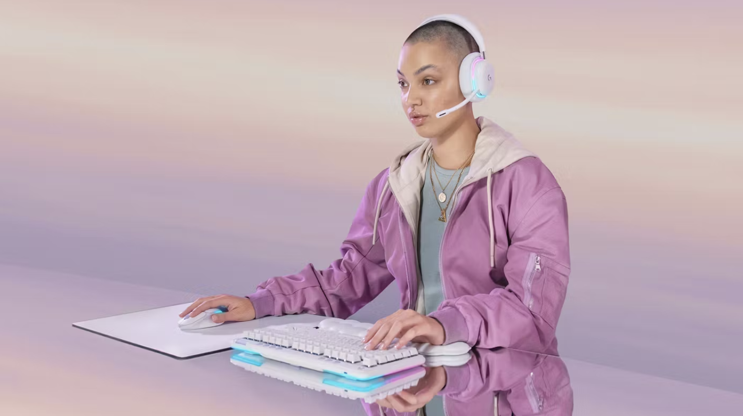 Young woman with shaved head wearing a pink jacket and headset, working on a computer with a keyboard and mouse on a reflective table against a pastel-colored background.