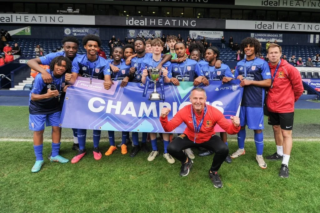 A youth soccer team in blue uniforms celebrating their championship victory on a soccer field. They are holding a large banner that says 'CHAMPIONS,' with a trophy in the middle, and are posing with a man in a red jacket.
