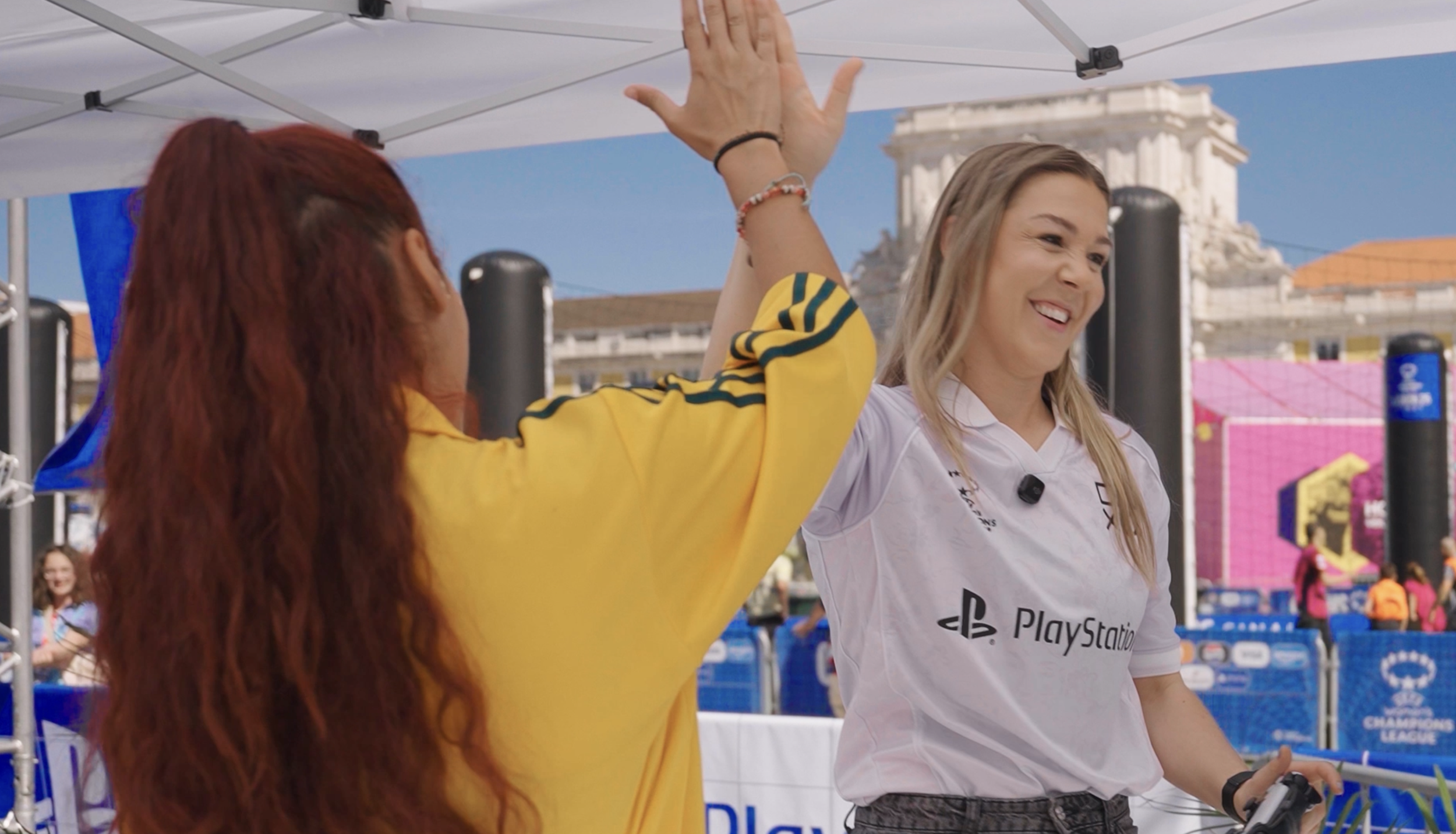 Two women giving a high five at an outdoor event with a PlayStation branding, crowd in background, and a historic building in the distance.