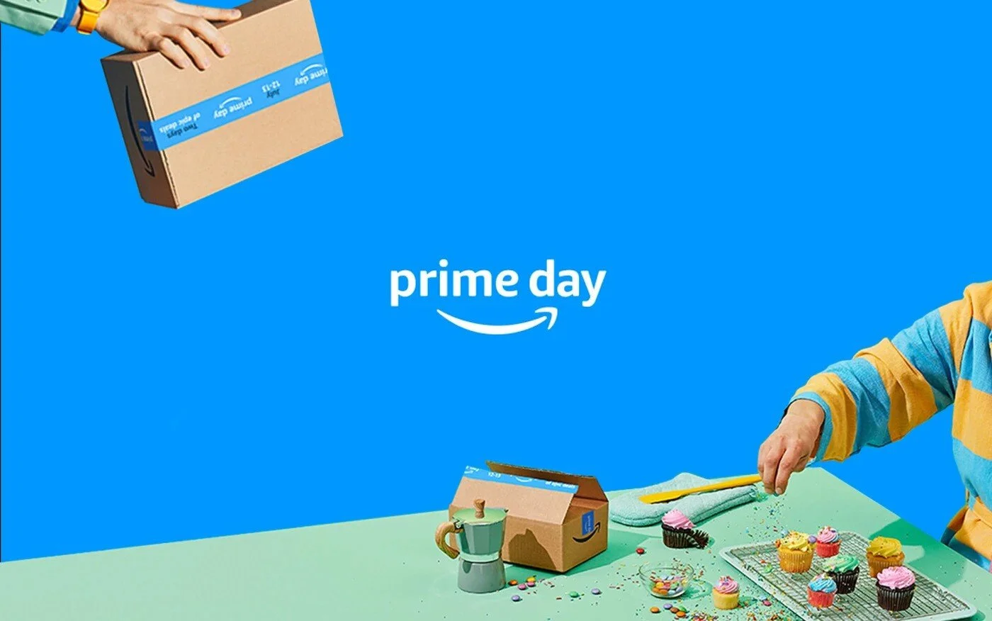 Amazon Prime Day advertisement featuring a blue background with the words "prime day" and a smile arrow. A person wearing a striped yellow and blue shirt is seen reaching for cupcakes on a table, with a brown Amazon Prime box, a coffee mug, and various colorful sprinkles and candies on the table.