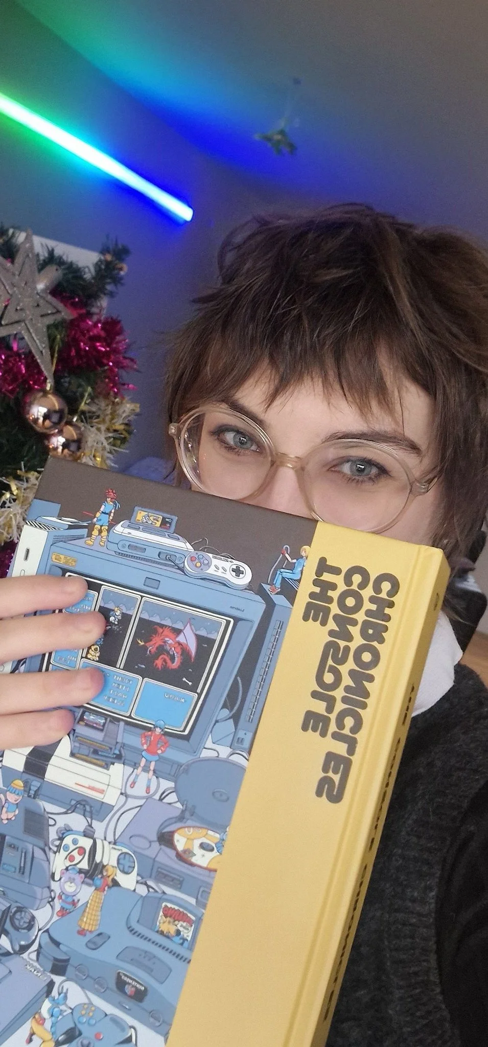 Person with short hair and glasses holding a boxed Sega Genesis video game console, with a Christmas tree in the background decorated with ornaments and tinsel, under colorful ceiling lights.