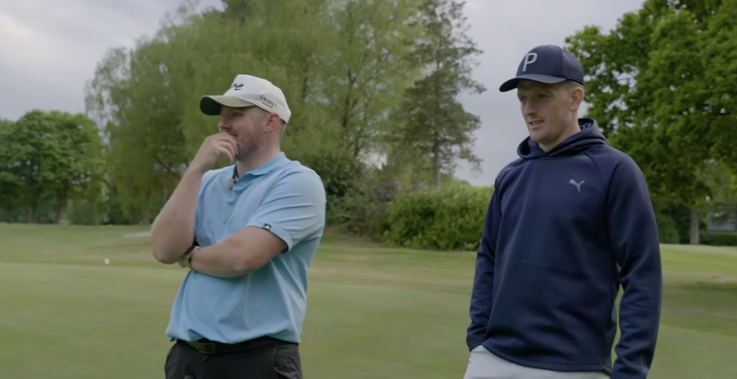 Two men standing on a golf course, one in a light blue polo and white cap, the other in a navy blue hoodie and navy cap, with trees and cloudy sky in the background.