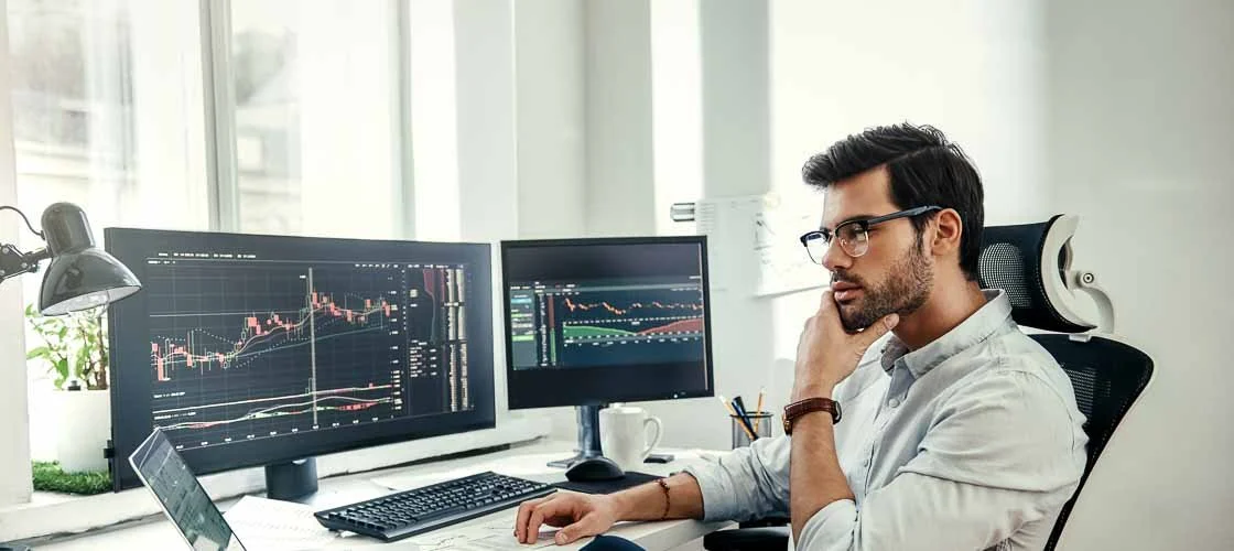 A man wearing glasses and a white shirt working at a desk with three computer monitors displaying stock trading charts.