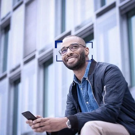 Smiling man wearing ZEISS Digital spectacle lenses, sitting outside holding a smartphone, in front of a modern glass building.