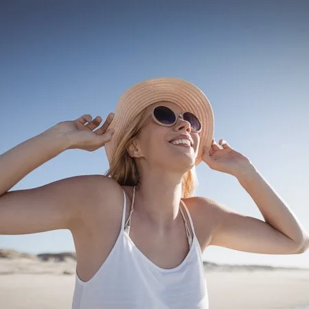 A woman wearing sunglasses and a wide-brimmed hat smiling at the beach.