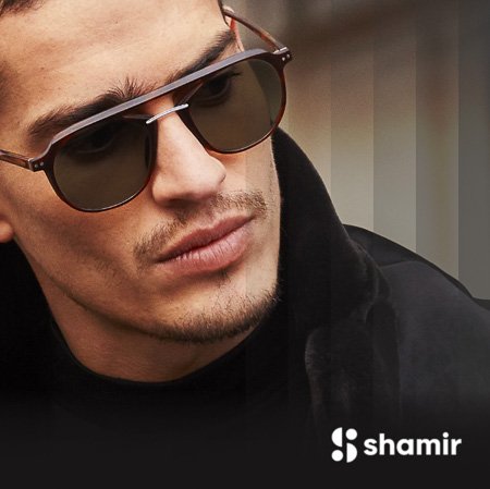 Close-up of a young man wearing sunglasses and a black jacket, with a blurred background, and the shamir logo in the bottom right corner.