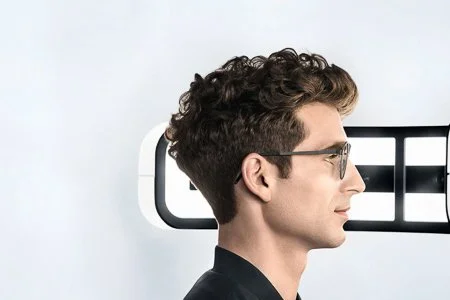 A man with curly hair wearing glasses looking to the right, with an eye examination machine behind him.