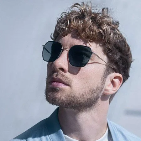 Young man with curly hair and beard wearing sunglasses and a light blue shirt.