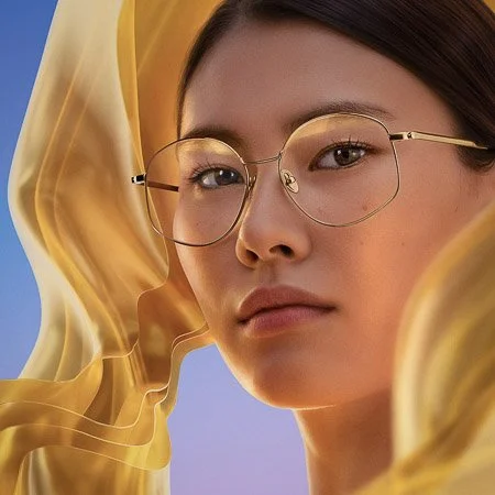 Close-up of a young woman with glasses, surrounded by flowing yellow fabric, against a solid blue background.