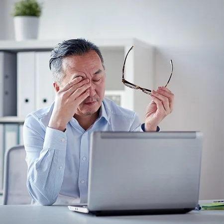 Man sitting at a desk, holding glasses in one hand and rubbing his temple with the other, in front of a laptop