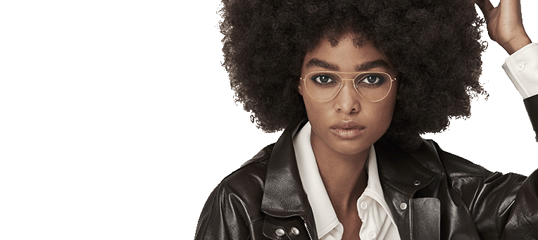 Young woman with curly hair and glasses wearing a leather jacket and white shirt, posing with her hand on her head.