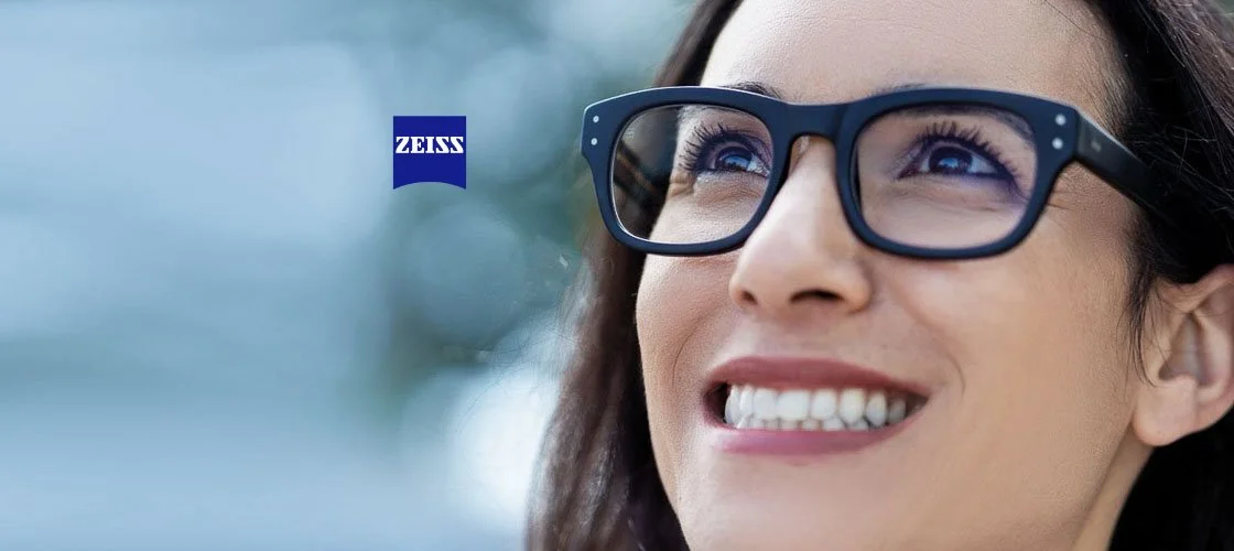 A woman wearing ZEISS spectacles outdoors, smiling and looking up, with the Zeiss logo visible in the background.