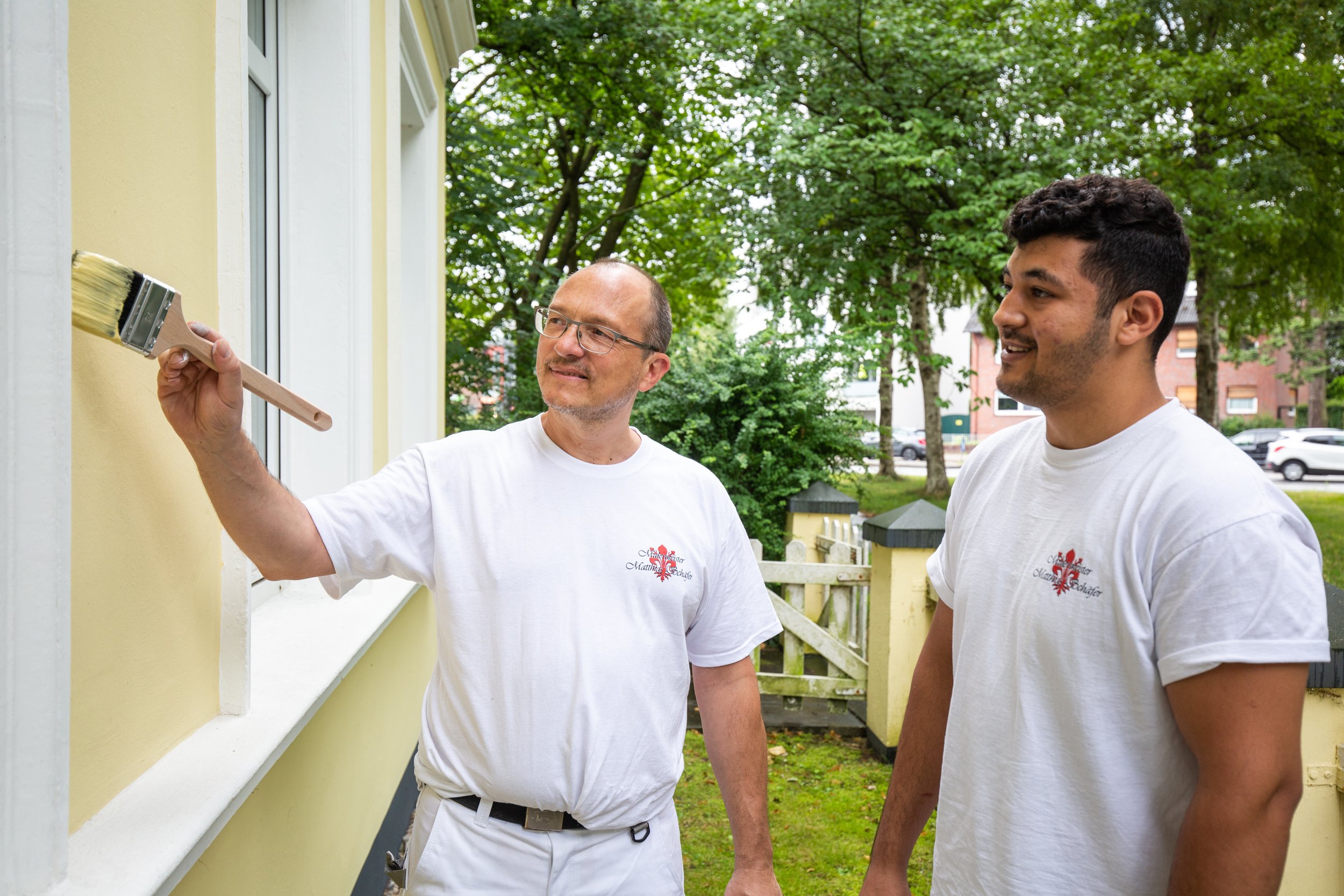 Zwei Männer in weißen T-Shirts stehen vor einer gelben Hauswand, während einer mit einem Pinsel die Wand streicht. Im Hintergrund sind Bäume und einige Häuser zu sehen.