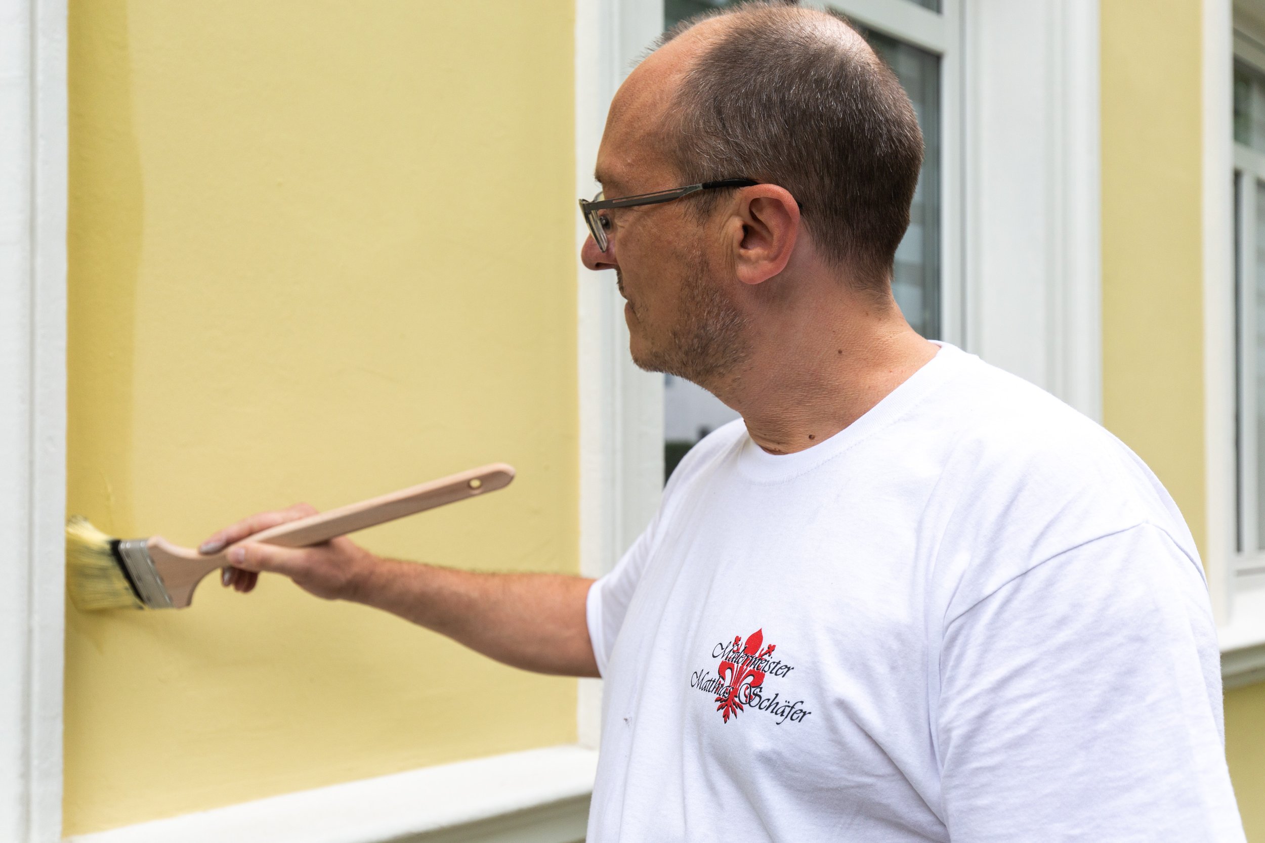 Ein Mann mit Brille streicht eine gelbe Wand mit einem Pinsel. Er trägt ein weißes T-Shirt.