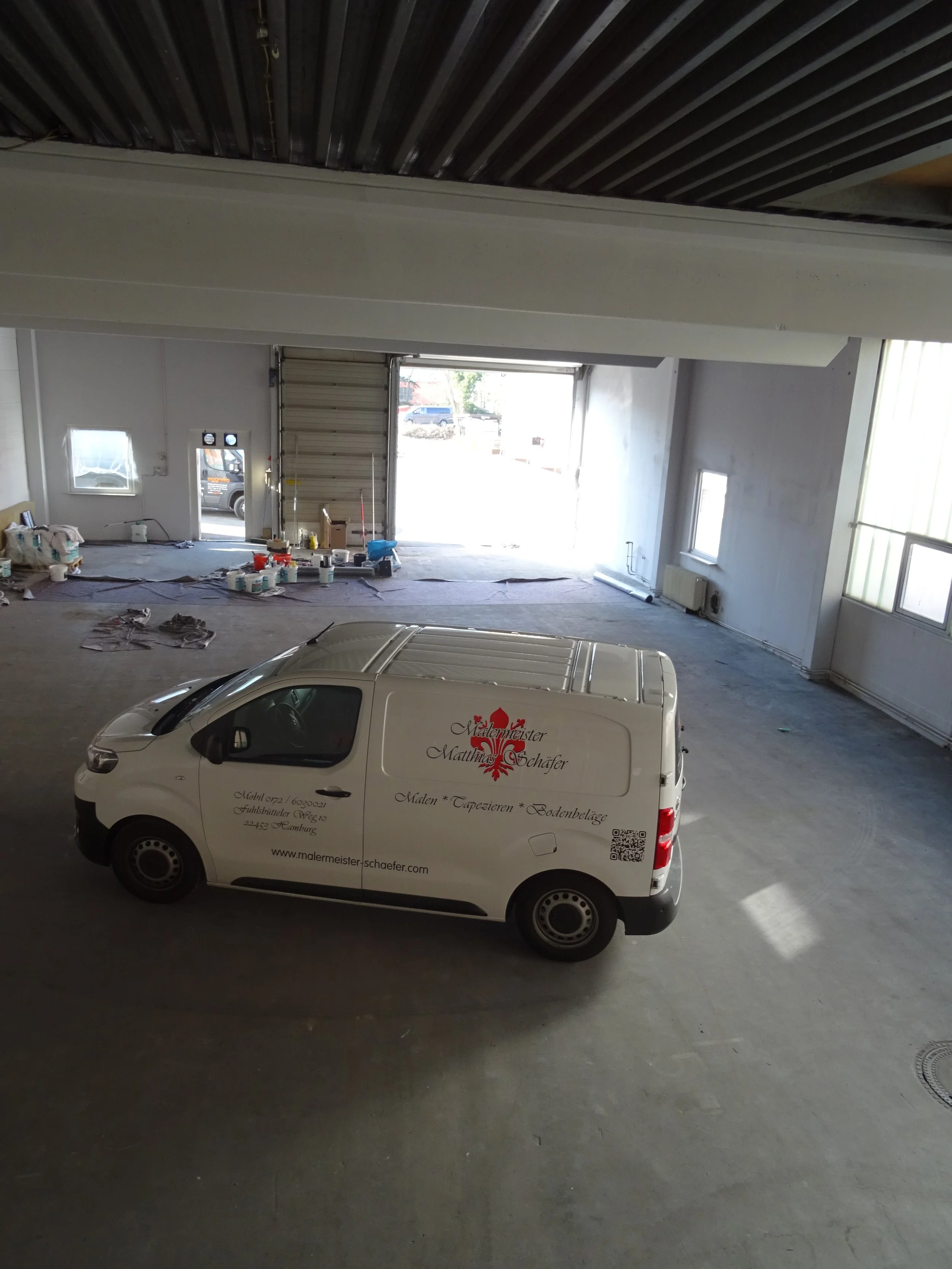 Innenraum einer Lagerhalle mit einem weißen Lieferwagen und Malerutensilien am Boden.