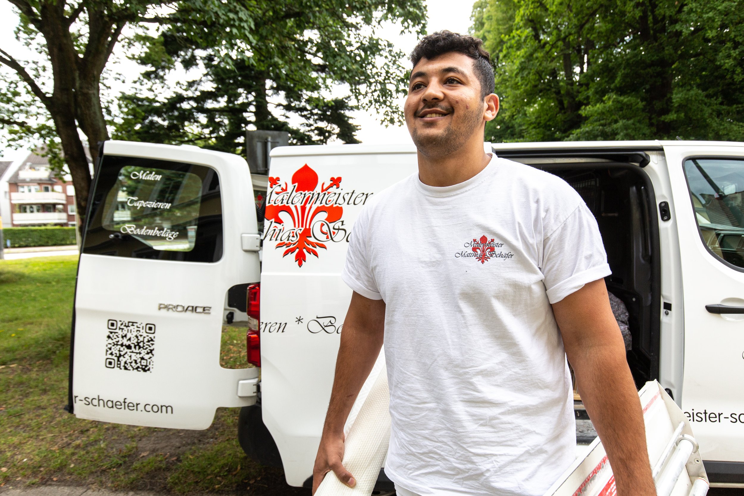 Ein Mann in einem weißen T-Shirt steht neben einem Lieferwagen mit der Aufschrift "Malen, Tapezieren, Bodenbeläge". Der Hintergrund zeigt Bäume und Wohngebäude.