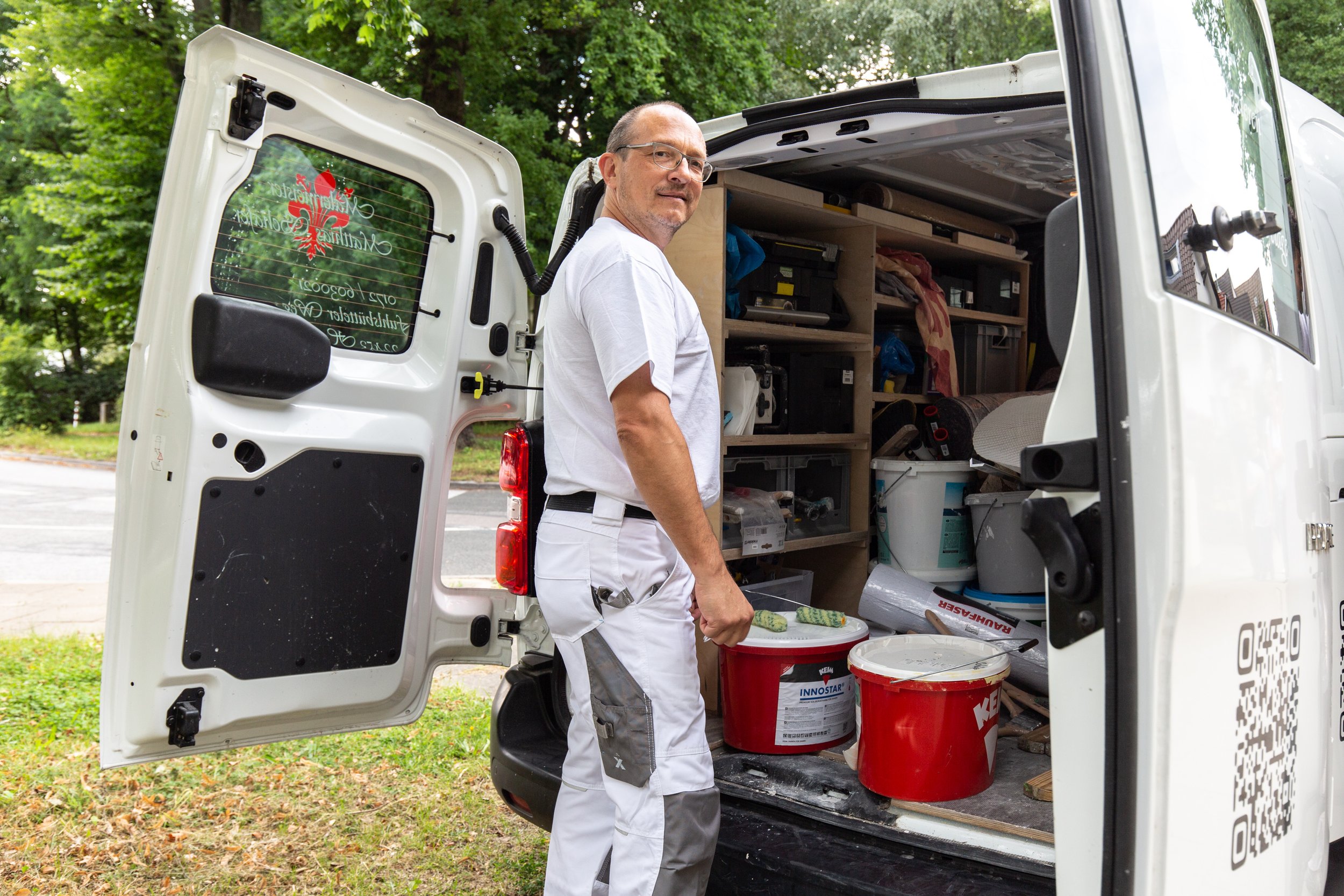 Ein Mann in Arbeitskleidung steht an einem geöffneten Transporter, der mit Werkzeugen und Farbeimern beladen ist, auf einer grünen Fläche neben einer Straße.