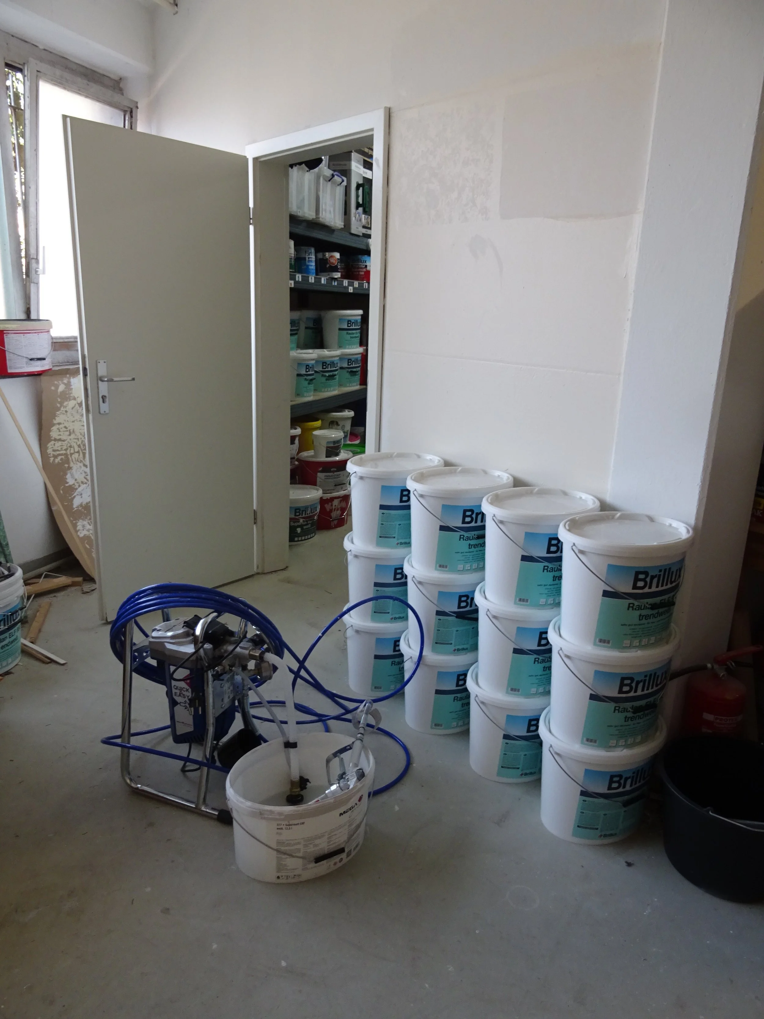 Lagerraum mit offenen Tür, Eimer mit Wandfarbe "Brillux" gestapelt, Farbspritzgerät und Eimer auf dem Boden.